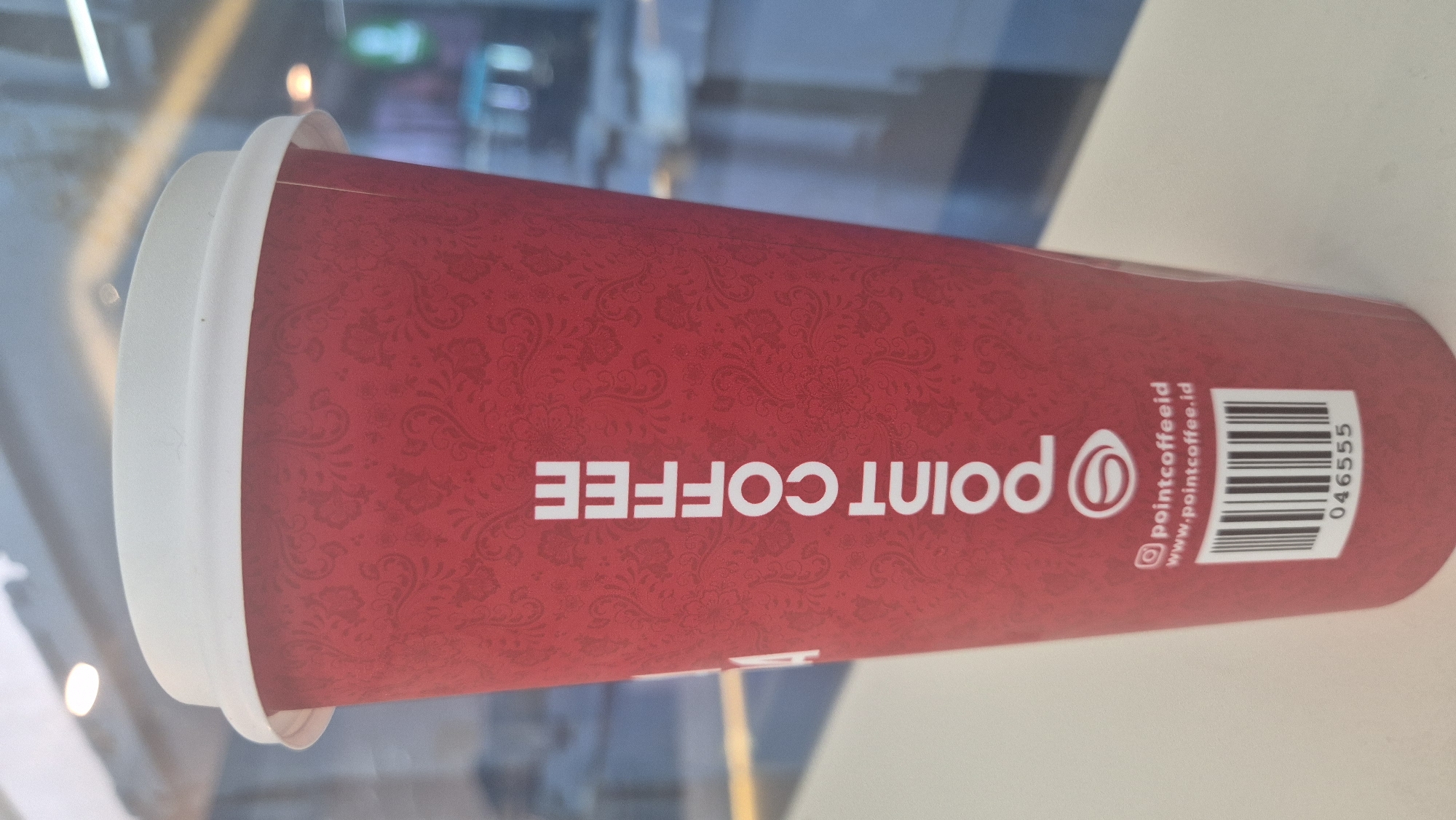 A close-up, vertical shot features a tall, red paper coffee cup with a white lid. The cup has a subtle, repeating paisley-like pattern in a slightly darker shade of red. The text "POINT COFFEE" is printed vertically in large, white sans-serif letters on the front of the cup. Below this text, there is a white rectangular area containing social media and website information, including "@pointcoffeeid" and "www.pointcoffee.id", followed by a barcode and the number "046555". The cup is placed on a white surface, likely a table, with a blurred background suggesting an indoor setting with windows and lights. The lighting is diffused, creating a soft glow on the cup and lid. The overall scene is clean and well-lit.