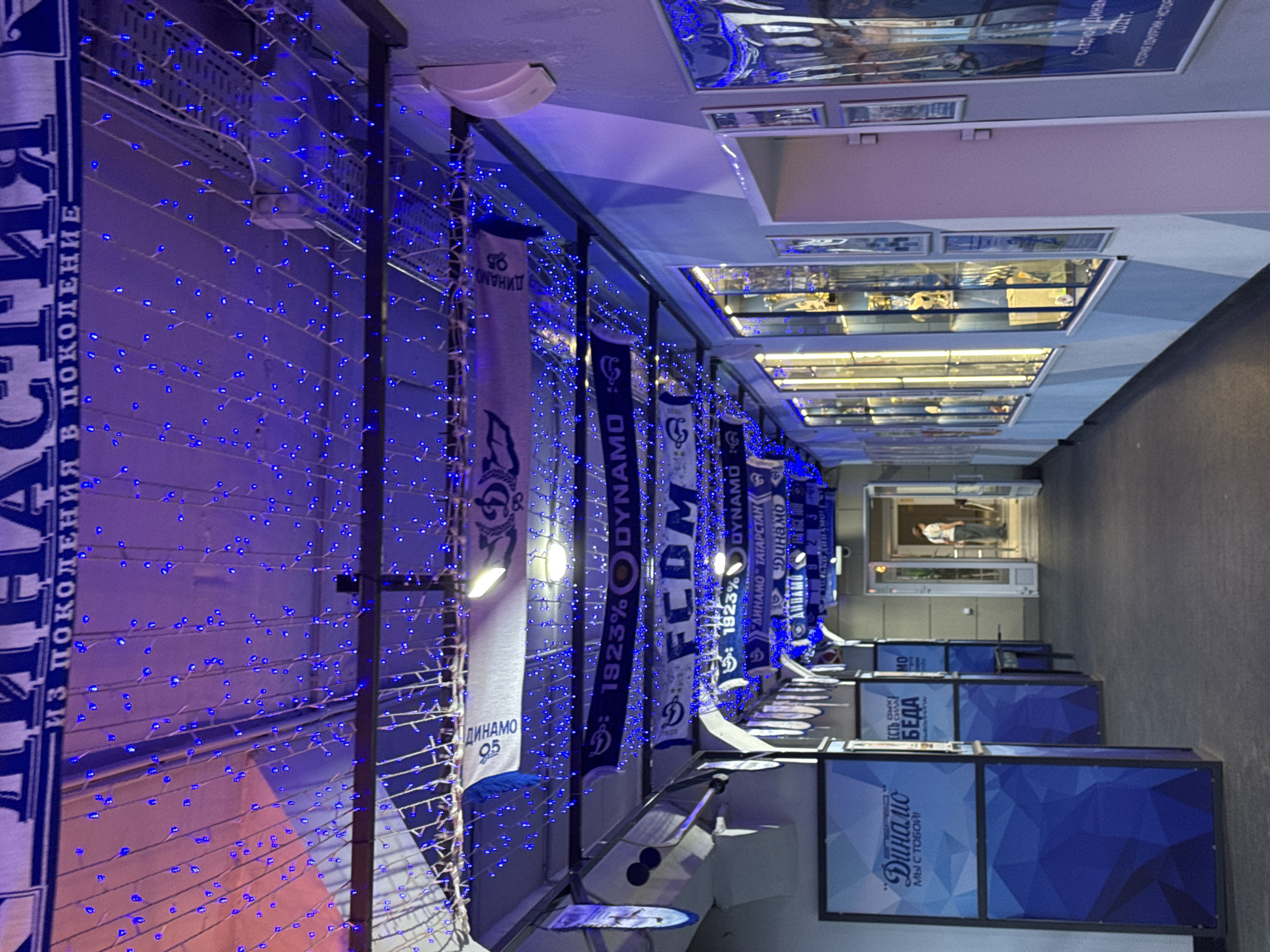 The image depicts a dimly lit hallway decorated with blue string lights and sports-themed banners. The hallway appears to be part of a museum or exhibition space dedicated to the Dynamo sports club, as evidenced by the prominent "Dynamo" branding on various banners and displays. On the left side of the hallway, there are large blue display panels with geometric patterns and text that reads "Динамо" (Dynamo) and "Мы с тобой!" (We are with you!). Further down the hallway, there are display cases filled with artifacts, likely trophies and memorabilia. On the right side, a person in a light-colored shirt and dark pants is walking away from the camera towards an open doorway. The overall ambiance is one of a sporting heritage exhibition, potentially located in Izhevsk, Russia, given the Dynamo club's presence there.