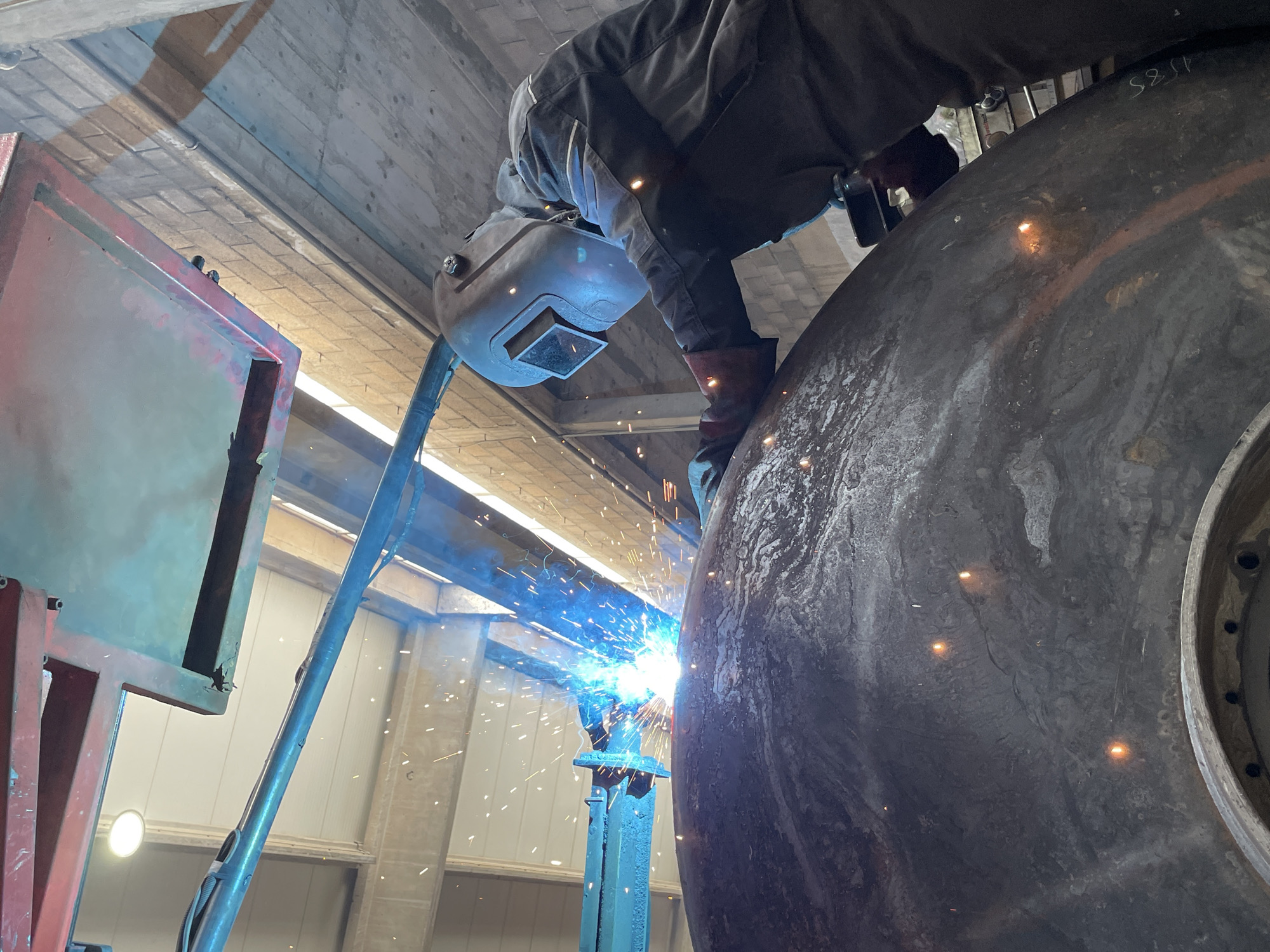 A welder is working on a large metal object, likely a wheel or tank, indoors in Čeluga, Montenegro. The welder wears protective gear, including a welding mask and gloves, and is positioned over the object. Sparks fly from the welding torch. A red structure is to the left of the welder. The environment appears to be an industrial building with concrete floors and ceilings.