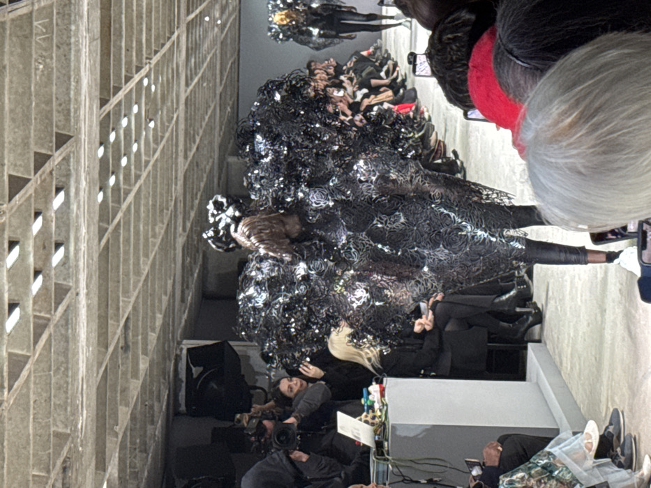 An indoor fashion runway show in Paris, France, specifically at the Carreau du Temple, is depicted. The scene features a central model walking away from the viewer on a light-colored runway. The model wears an expansive, voluminous, sculptural garment composed of numerous reflective, dark, crumpled elements resembling petals or abstract organic forms, covering the torso and shoulders. This piece is paired with black trousers and white sneakers. The model's hair is dark and slicked back, topped with a dark, intricate headpiece that complements the main garment.
Further down the runway, a second model in a similar, though less defined, black sculptural outfit is visible. On both sides of the runway, rows of spectators are present, with several individuals in the foreground and midground holding smartphones to record the event. To the immediate left of the runway, a group of media personnel is positioned, including an individual operating a professional video camera and another near a grey podium with a laptop and other equipment. The venue features a distinctive concrete waffle slab ceiling with numerous recessed rectangular light fixtures providing illumination. The walls appear to be a light grey or concrete, and the floor of the runway is a smooth, light surface.