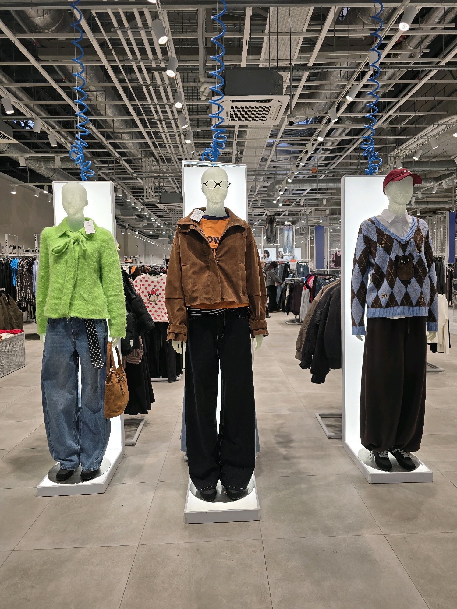 Three mannequins are displayed in a retail store, likely a clothing store, in Khimki, Russia. The mannequins are arranged in a row from left to right, each on a white platform.
The leftmost mannequin is wearing a bright green, fuzzy collared sweater with a bow tie detail, light blue wide-leg jeans, a black belt with a white polka dot pattern, and black shoes. A brown suede handbag is hanging from its right side.
The center mannequin is dressed in an orange t-shirt with an "OW" graphic, a brown suede-like jacket, and black wide-leg pants, paired with black flat shoes. It is also wearing glasses.
The rightmost mannequin is sporting a white collared shirt under a blue and brown argyle patterned sweater with a teddy bear motif. It is wearing dark brown wide-leg pants and black sneakers, with a red baseball cap.
The background reveals a well-lit store interior with industrial-style ceiling infrastructure, including pipes, conduits, and exposed wiring. Blue coiled cords hang prominently from the ceiling, adding a decorative element. Racks of clothing are visible behind the mannequins, suggesting a typical retail display. The floor is tiled with large, light-colored square tiles. The overall impression is a modern, somewhat industrial retail environment.