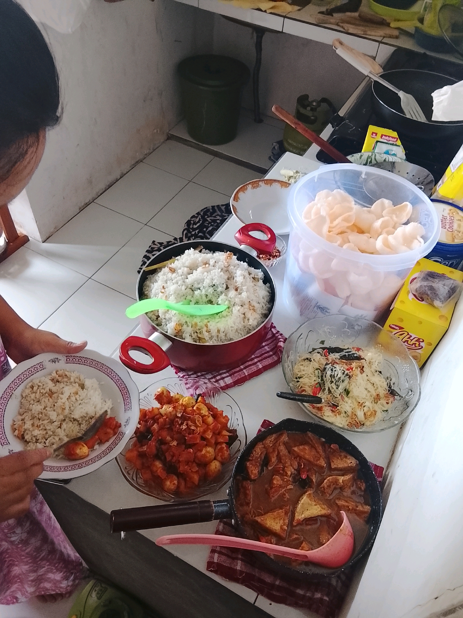 In Kucur, Indonesia, a person's arm is visible on the left side of the image, serving a plate of rice. The scene is a kitchen with a tiled floor and white walls. Several dishes are arranged on a white countertop. A large pot of white rice is in the center, with a green serving spoon. Next to it, a clear plastic container holds a pile of light-colored crackers. A glass bowl contains a noodle dish with vegetables. Another glass bowl holds small red items, likely quail eggs in sauce. In the foreground, a black pan filled with triangular pieces of tofu in a dark sauce is visible, with a pink ladle resting in it. A yellow box, possibly for snacks, and a container of "butter cookies" are also on the counter. In the background, a green trash can and kitchen appliances are present.