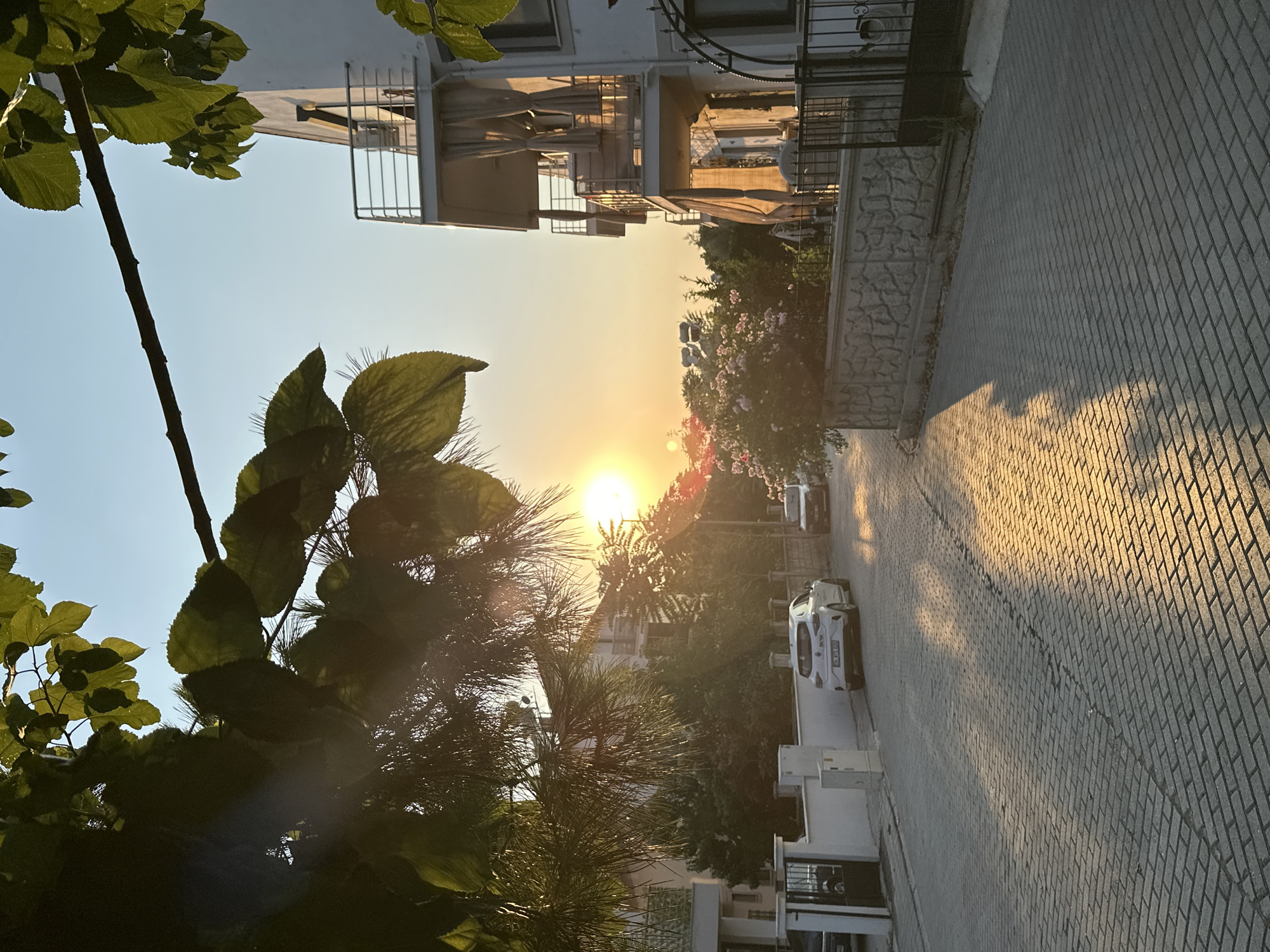 The image depicts a street scene in Fethiye, Turkey, during sunset. The foreground shows a cobblestone street with shadows cast by overhanging trees and a building. Two white cars are parked on the side of the street. A multi-story building with balconies and curtains is visible on the right side of the image, bathed in the warm light of the setting sun. Lush green trees and a bush with pink flowers are present, contributing to the residential feel of the area. The sky is clear with a gradient of orange and yellow hues from the sunset. There are no people visible in the frame.