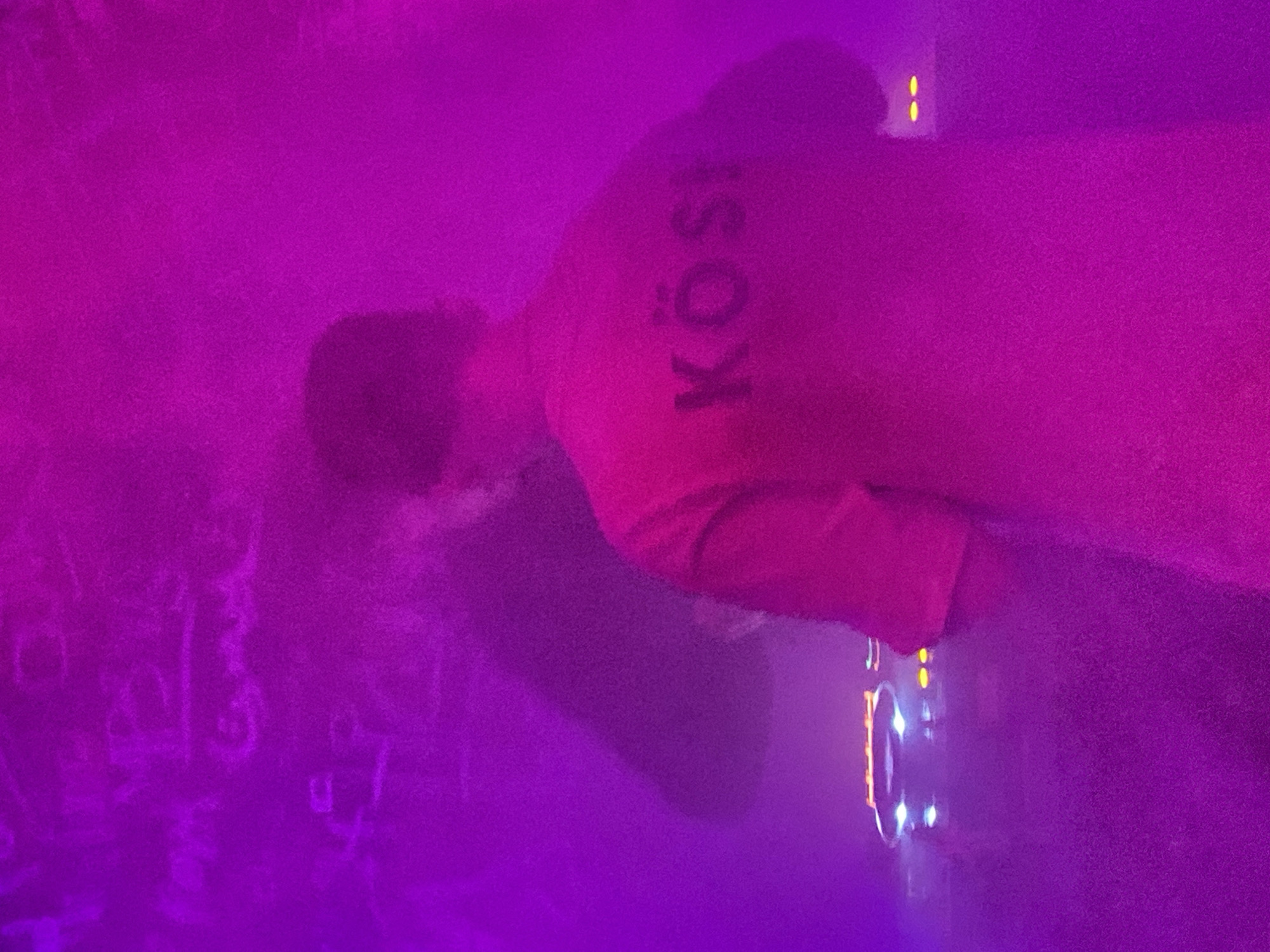 A person wearing a shirt with the letters "KÖS" printed on the back stands facing a mixer. The setting is dimly lit, with purple and pink hues dominating the scene. The location is potentially Шебунино, Russia.