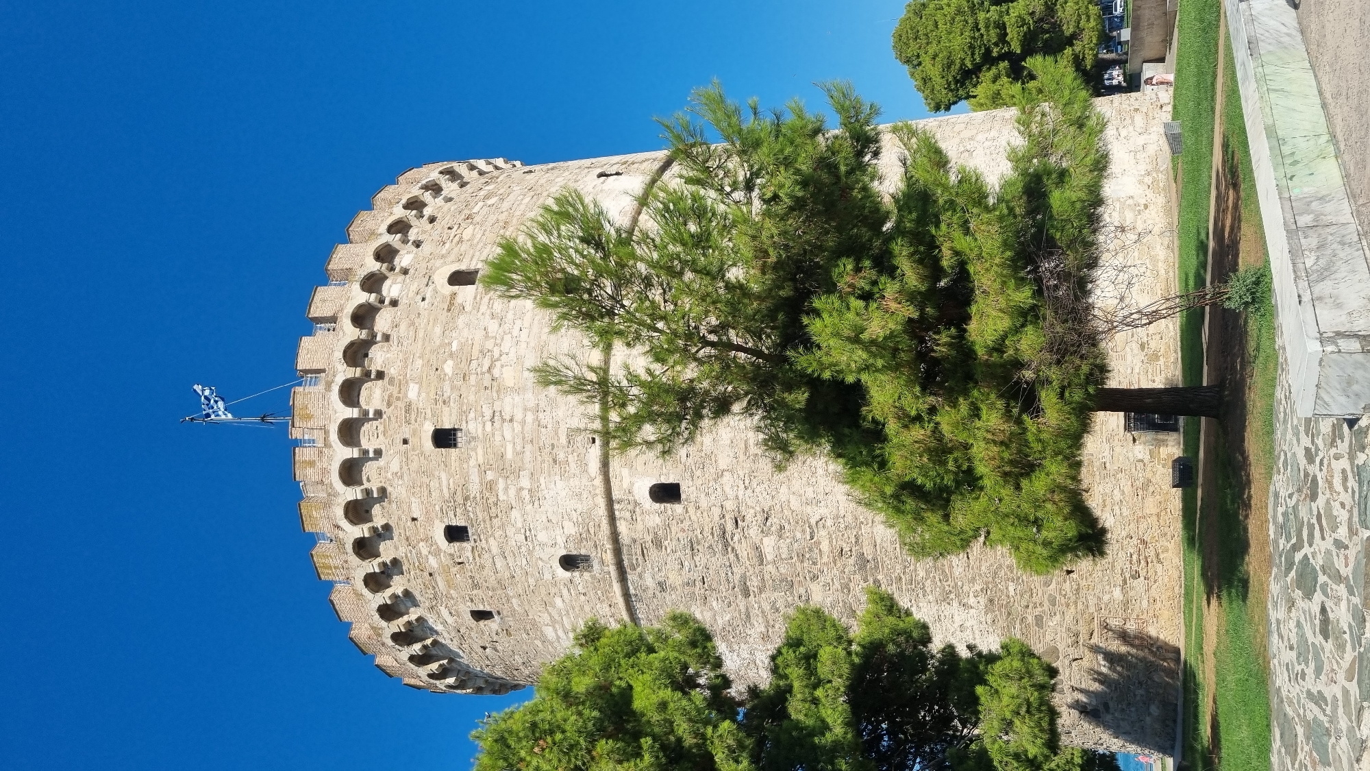 The White Tower, a landmark in Thessaloniki, Greece, stands tall against a clear blue sky. This cylindrical stone tower features a crenellated parapet and numerous small windows. A Greek flag flies from a flagpole at its summit. Lush green pine trees partially obscure the tower, with their branches casting shadows on its ancient stone facade. The ground in front of the tower is covered in grass and a stone-paved area. In the distance, a few people are visible, along with parked cars and other trees, suggesting a public park or promenade setting within the city. The overall scene depicts a sunny day in a historical urban landscape.