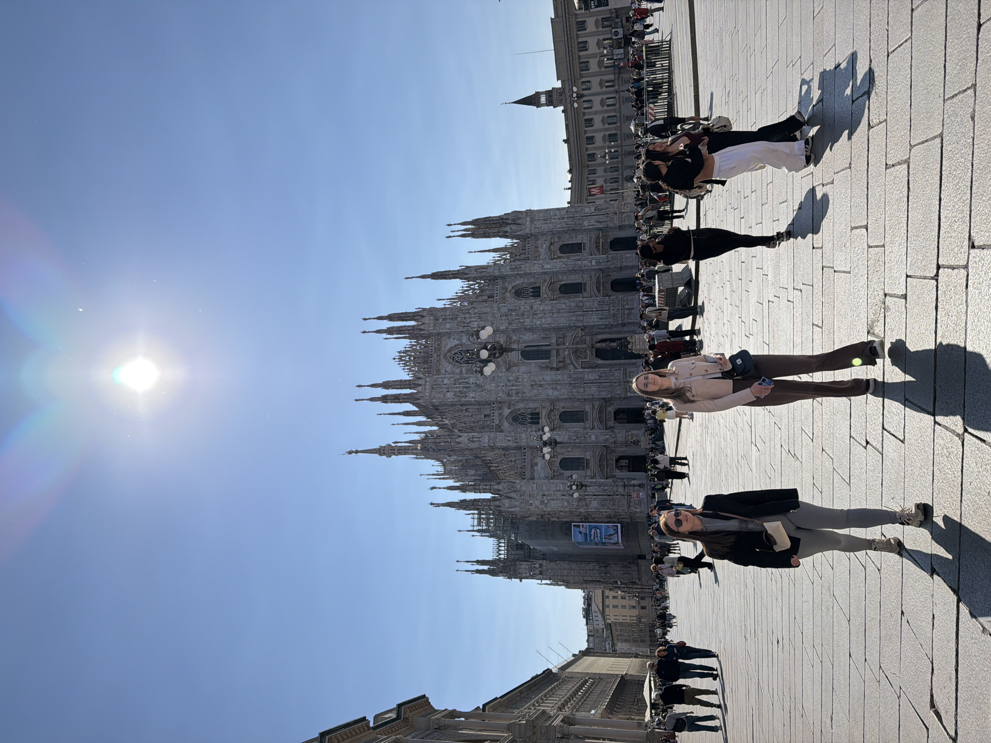 A wide-angle, slightly low-angle photograph captures a bustling public square on a bright, sunny day, dominated by the majestic Duomo di Milano. The grand Gothic cathedral, with its intricate spires, detailed facade, and statues, stands prominently in the mid-ground. Scaffolding is visible on a section of the Duomo's exterior, suggesting ongoing restoration or maintenance work, with a large poster affixed to it.
The sky is a clear, brilliant blue, with the sun intensely shining from the upper left, causing a distinct lens flare with rainbow-colored streaks. This strong sunlight casts long, sharp shadows of people and structures across the light-colored paved square, indicating either early morning or late afternoon.
Numerous people are scattered across the square, appearing as visitors or tourists. Two women are prominent in the central foreground, both looking towards the camera. One wears a light-colored jacket over dark pants, and the other, slightly behind her, wears a black jacket, grey pants, and sunglasses. Other individuals and small groups are seen walking, standing, and observing the iconic landmark.
In the background and to the right, other historic city buildings flank the square. One building displays a red "M" sign, likely indicating a metro station entrance. Barriers or railings are also visible along the edges of the paved area. The image conveys a lively atmosphere in a major tourist destination in Milan, Italy.