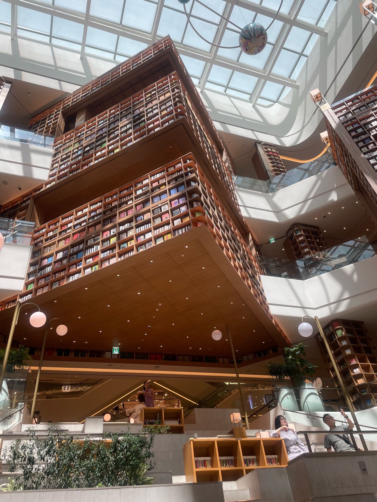 The image shows an interior view of a multi-level bookstore, likely Starfield Library, located in the Starfield COEX Mall in Gangnam-gu, Seoul, South Korea. The architecture is characterized by towering wooden bookshelves that ascend multiple floors, creating a dramatic and cavernous space. A large, translucent skylight system covers the ceiling, allowing natural light to filter in and casting geometric shadows. A colorful spherical art installation hangs from the ceiling.
In the foreground, a woman in a light purple dress is seated on a tiered platform, looking towards a man holding a phone up, possibly taking a photograph. They are near a low wooden bookshelf containing rows of books. Green plants are also visible on the platforms and in planters.
On higher levels, more bookshelves are filled with books, and escalators are visible, suggesting the multi-story nature of the space. People can be seen on different levels, browsing or walking. An emergency exit sign with a green figure is visible on one of the walls. The overall ambiance is one of modern architectural design combined with a vast collection of literature.