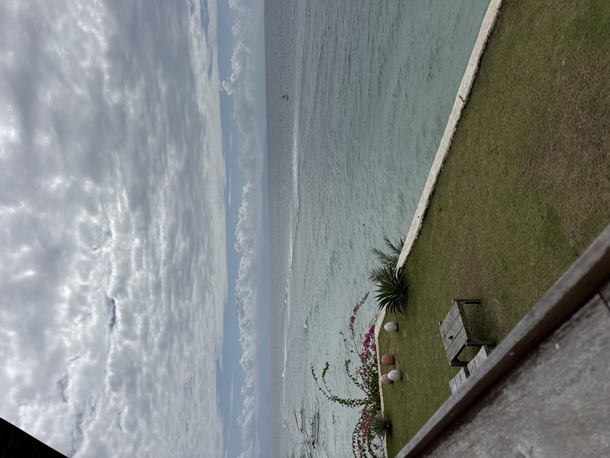 The image depicts a serene seascape, likely from a resort or vantage point in Nusa Penida, Indonesia. The foreground features a neatly manicured grassy area with a simple wooden table and bench set. On the grass, several decorative spherical objects, some with sculpted faces, are arranged. A lush green plant and flowering vines with pink blossoms add a touch of color to the scene. Beyond the lawn, a clear turquoise ocean stretches to the horizon, meeting a sky filled with dramatic, textured clouds. In the distance, the silhouettes of mountains are visible. Several small boats are anchored near the shore, and a few individuals can be seen on the water, possibly enjoying water sports or fishing. The overall atmosphere is tranquil and picturesque, capturing the beauty of a tropical coastal landscape.