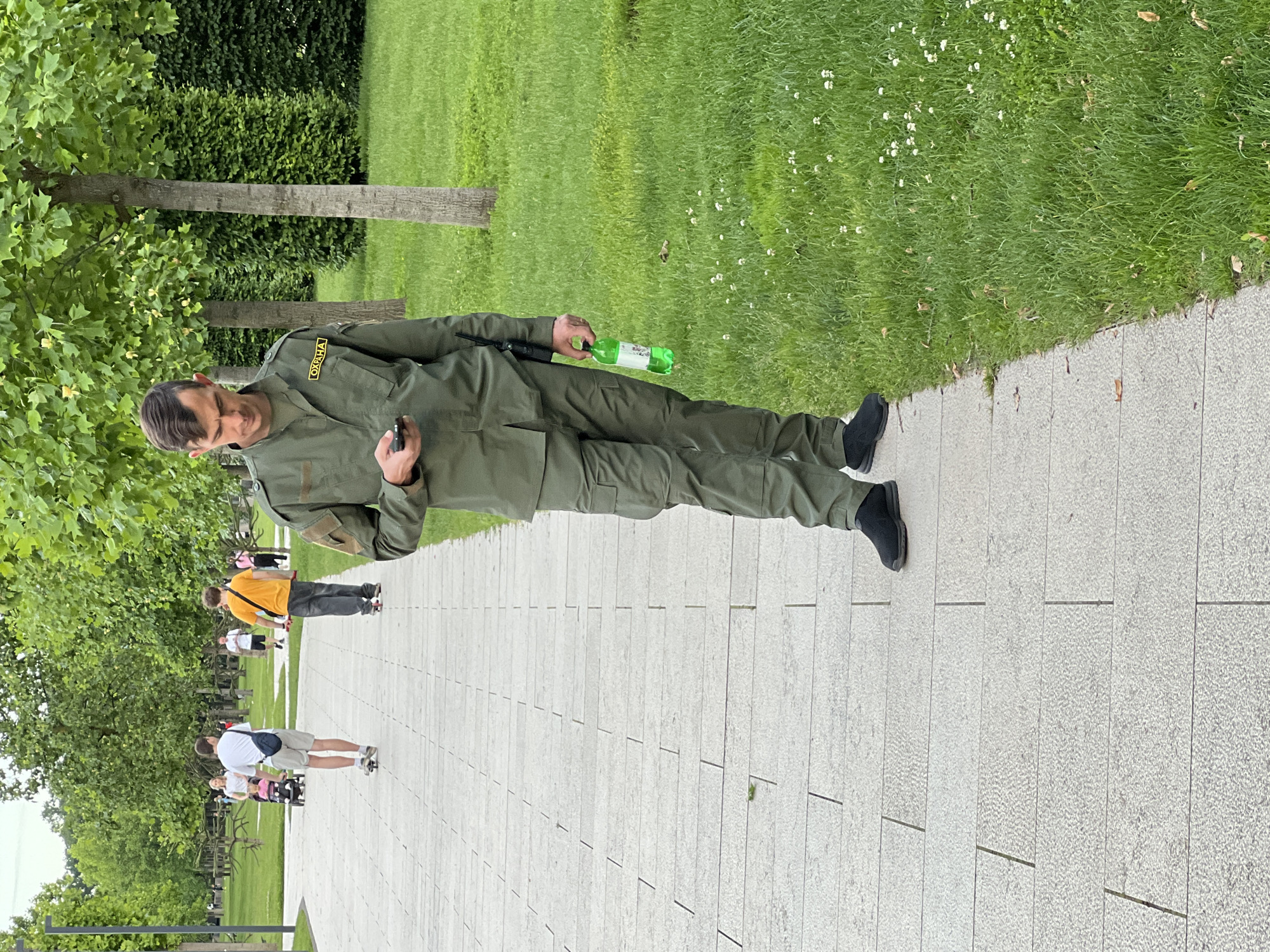 This media file captures a male security guard in a park-like setting in Krasnodar, Russia, during daytime. The image is oriented vertically (sideways), but depicts the scene as if the man is standing upright on a paved path.
The main subject is a man wearing a dark olive green uniform, which appears to be a two-piece set or a jumpsuit, featuring chest and arm pockets. He has dark hair, styled with a part, and is wearing glasses. On his right shoulder, a yellow patch with black Cyrillic text clearly reads "ОХРАНА" (OKHRANA), which translates to "SECURITY" or "GUARD". He holds a small, dark object, possibly a mobile phone or a communication device, in his left hand, and a green plastic bottle with an indecipherable white label in his right hand. A dark, slender object, possibly a radio or a weapon component, is visible tucked into his side. His expression is neutral to slightly serious, consistent with someone on duty.
He stands on a wide pathway made of light-grey, rectangular paving stones. To his left, a lush green lawn extends, dotted with small white flowers. Beyond the lawn are several trees with visible trunks and vibrant green foliage, and a dense green hedge. To his right, more trees and greenery frame the path.
In the background, further down the paved path, several other people are visible, casually walking. One person wears a bright orange t-shirt and dark pants, while another wears a white t-shirt, shorts, and carries a dark blue shoulder bag.
The scene suggests a pleasant day, with bright, even lighting indicating it's daytime. The overall setting is a well-maintained public park or a similar outdoor recreational area. The man's uniform and the "ОХРАНА" patch confirm his role as a security guard, consistent with the provided location context of Krasnodar, Russia.
