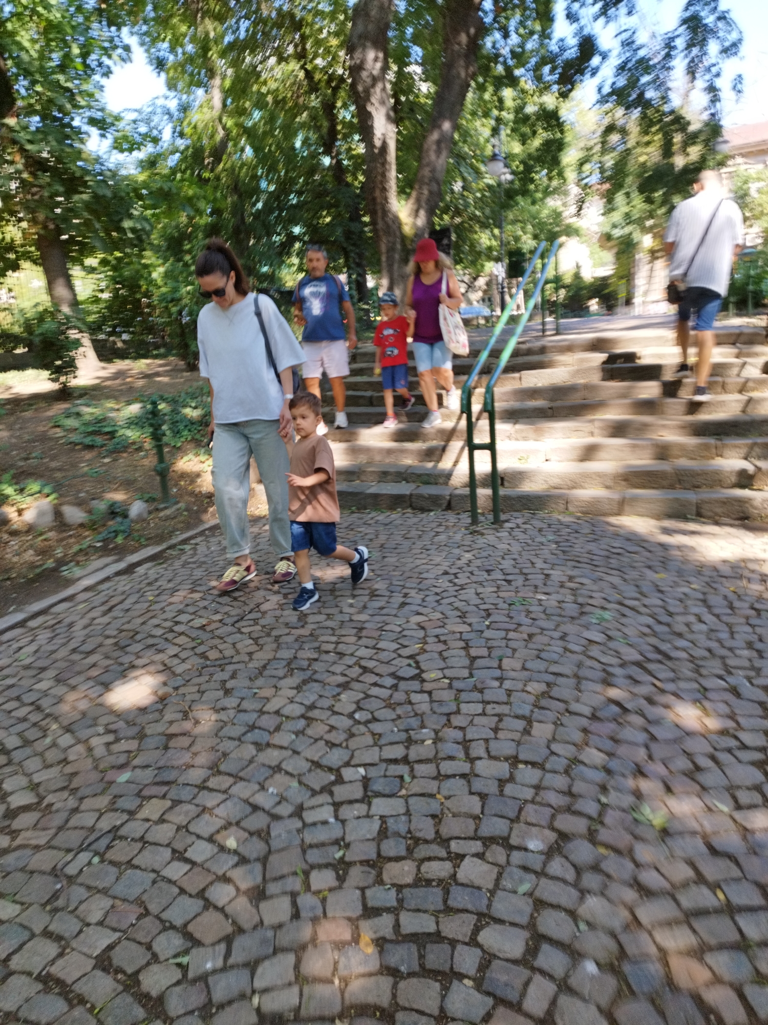 A medium shot captures a group of people walking down a cobblestone pathway beside a set of stone stairs in a park setting, likely in Bucharest, Romania, given the style of the cobblestones and the general ambiance. The image has a slight blur, indicating motion.
In the foreground, a young boy with brown hair, wearing a tan t-shirt and dark blue shorts, is running down the pathway, his right hand extended forward as if reaching for something or indicating a direction. He is accompanied by a woman with dark hair pulled back in a ponytail, wearing a light-colored oversized t-shirt and grey trousers. She holds the boy's hand and walks beside him.
Further up the pathway and on the stairs, other individuals are visible. A man in a blue t-shirt and white shorts is walking up the stairs, carrying a backpack. A woman in a purple tank top and light blue shorts, wearing a red hat, walks down the stairs, holding a patterned tote bag. A young boy in a red t-shirt and blue shorts is walking alongside her on the stairs. A man in a white t-shirt and blue shorts is walking up the stairs further in the background.
The environment is lush with green trees and foliage, and sunlight filters through the leaves, creating dappled patterns on the ground. A green metal handrail runs alongside the stairs. The pathway is made of dark, uneven cobblestones. The overall impression is a family outing or a group enjoying a stroll in a park.