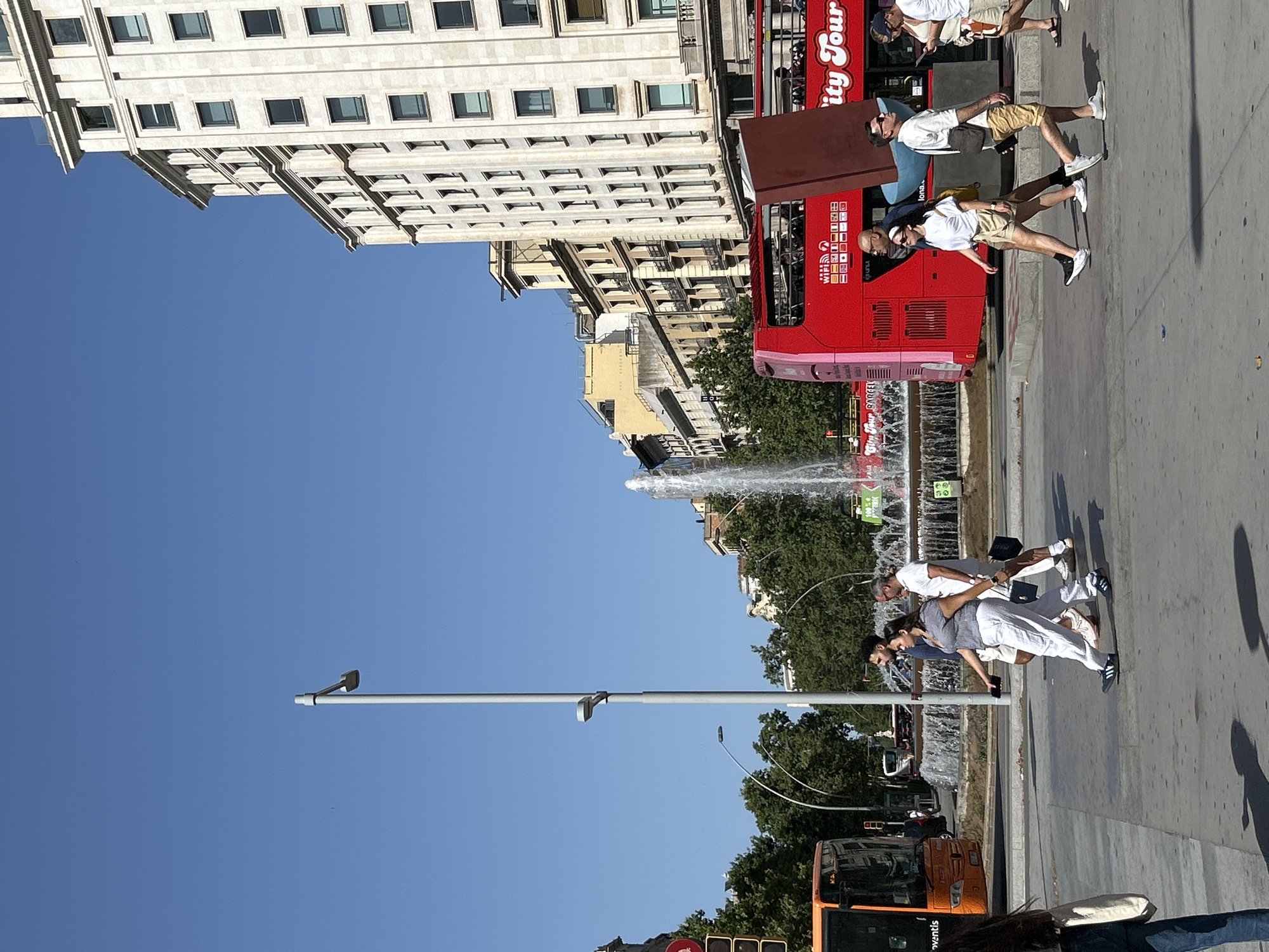 This media file captures a sunny day in Barcelona, Spain, likely in the late morning or early afternoon given the bright sunlight and clear blue sky. The scene is a busy urban street with a prominent red double-decker tour bus, characteristic of those found in many major cities, including Barcelona. The bus has "City Tour" text visible on its side, indicating its purpose.
Several people are visible in the image. Some are walking on the sidewalk, while others appear to be interacting with each other near the tour bus. One group of three individuals, two women and one man, are dressed in light-colored clothing, including white pants and tops, suggesting warm weather. They are posed in what appears to be a group photo or are engaged in a lighthearted activity. Another man, wearing a white t-shirt and beige shorts, is also visible, possibly a tourist.
In the background, a multi-story building with numerous windows lines the street, indicating a commercial or residential area. Adjacent to the tour bus, a fountain is active, adding a refreshing element to the urban landscape. Further down the street, an orange bus is also present, along with traffic lights and street lamps. The overall impression is of a vibrant, active city scene on a pleasant day.