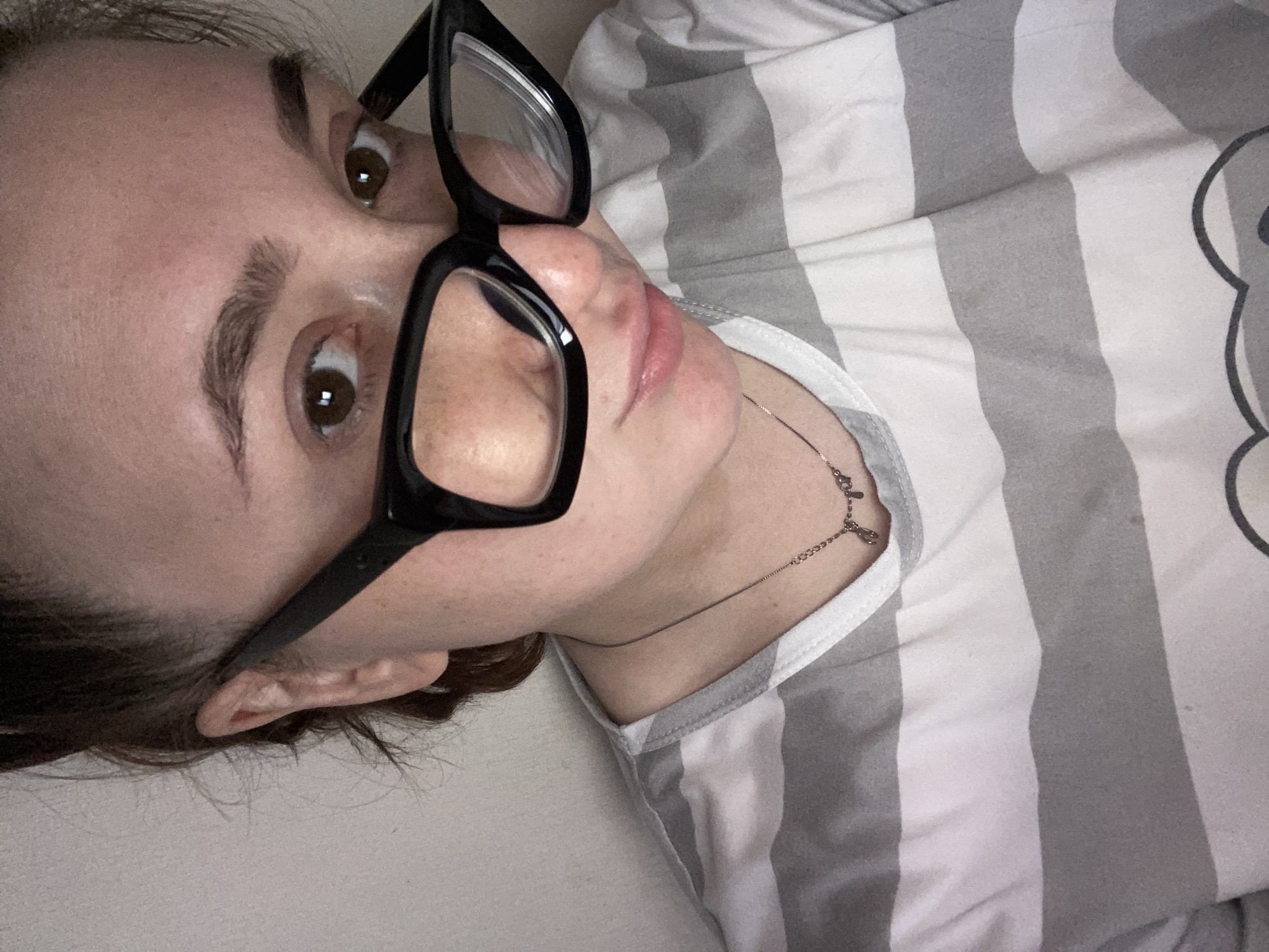 A woman with brown hair and eyes is wearing black-framed glasses and a striped gray and white shirt. She is lying down. A thin silver necklace is visible around her neck. The location is unknown and cannot be determined from the image. The city of Chaikovsky, Russia cannot be determined.