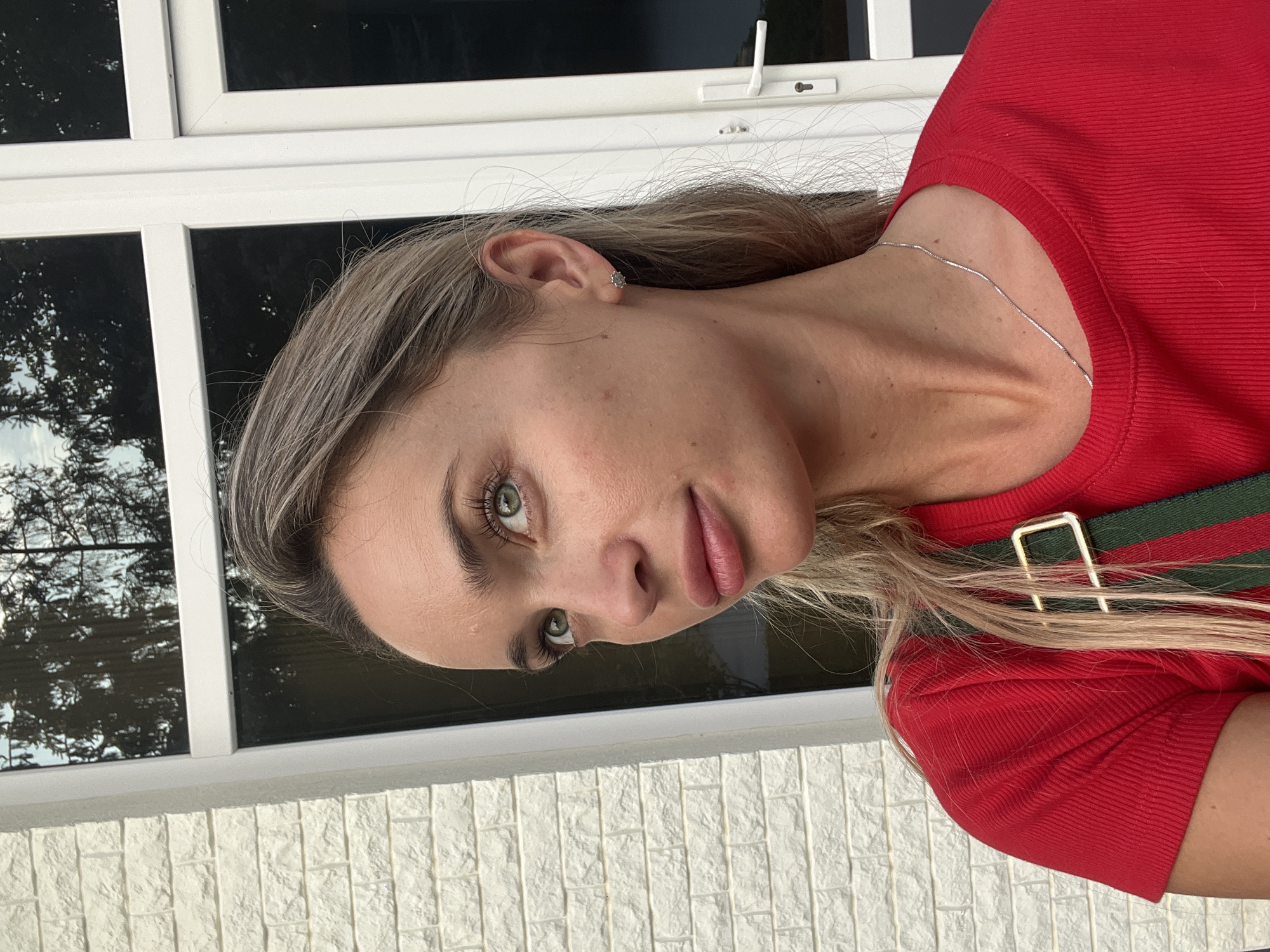 A young Caucasian woman with blonde hair and green eyes is shown in a close-up, upper-body portrait. She is wearing a red, ribbed t-shirt and a silver chain necklace. A green and red striped strap with a gold buckle is visible across her chest. She is wearing a small, star-shaped earring in her left ear. The woman is looking to her left, with her head slightly tilted. Her skin appears slightly flushed. The background consists of a white brick wall on the left and a white-framed window with dark panes on the right. Trees are reflected in the window. The setting is outdoors.