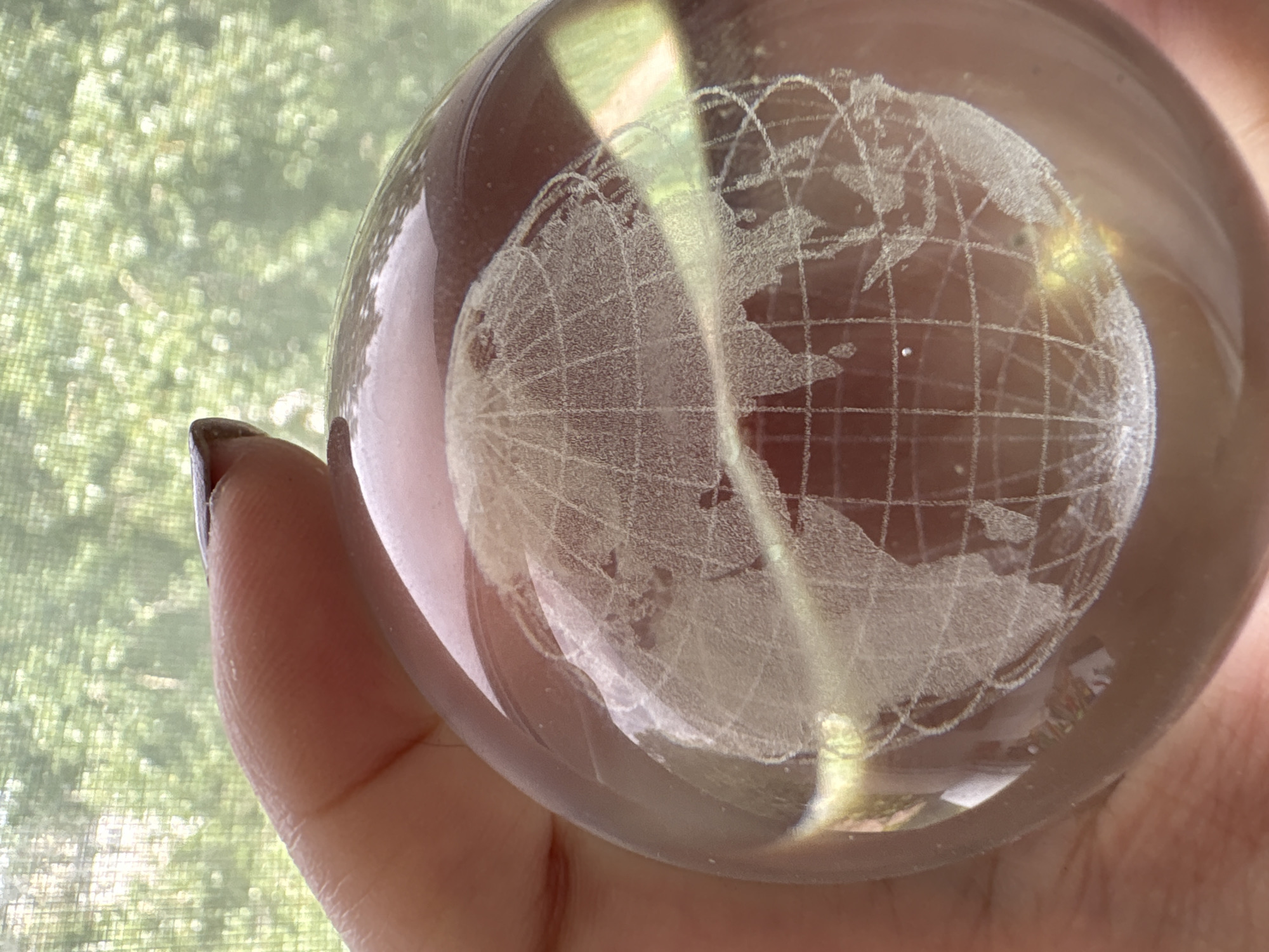 A hand is holding a transparent glass globe with a frosted etching of the Earth's continents and latitudinal/longitudinal lines. The background shows blurry greenery visible through a window. There is no indication of Izhevsk, Russia in the image.