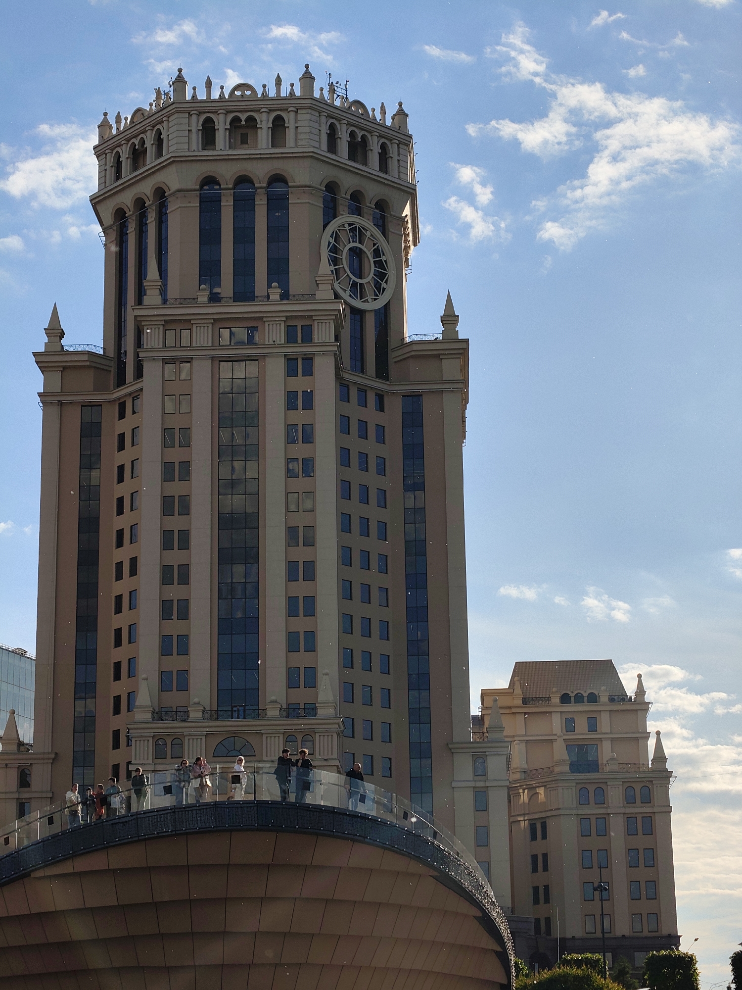 The image shows a tall building complex in Moscow, Russia. The primary building is a tan skyscraper with a distinctive octagonal tower at the top, featuring arched windows and a large, round decorative element resembling a clock face. The building's facade consists of vertical sections of windows and light-colored panels. In the foreground, a curved pedestrian bridge made of brown panels and glass railings is visible. A group of people are standing on the bridge, observing the surroundings. Another, smaller building with a similar architectural style is visible to the right of the main skyscraper. The sky above is blue with scattered white clouds.