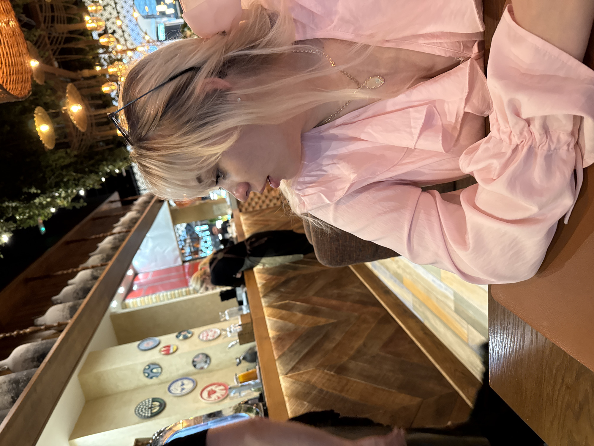 The image, taken in Perm, Russia, captures an indoor scene, likely a restaurant or cafe, during the evening. The main subject is a person with blonde hair, seen in profile, looking downwards with a focused expression. They are wearing a light pink, possibly satin, long-sleeved top with gathered cuffs and a gold necklace with a light-colored oval pendant. Glasses are resting on top of their head.
The person is seated at a wooden table featuring a herringbone pattern, next to a dark brown, leather-like seating surface. The background showcases the establishment's warm and inviting decor, characterized by numerous artificial light sources. String lights are visible in the upper left, alongside hanging light fixtures with woven or metallic shades, casting a soft, yellow glow.
Decorative elements include a ledge or shelf adorned with various circular plates or coasters displaying diverse patterns. Notable among these are a red plate with a white letter "P" and a blue plate with a white letter "E". Glassware and bottles are also present on this shelf. To the left, a wooden structure, possibly a railing or wall accent, is decorated with white, snow-like caps. Another person is partially visible, blurred, in the far background, seated at a different table. The overall scene suggests a cozy and modern dining or social atmosphere at night.