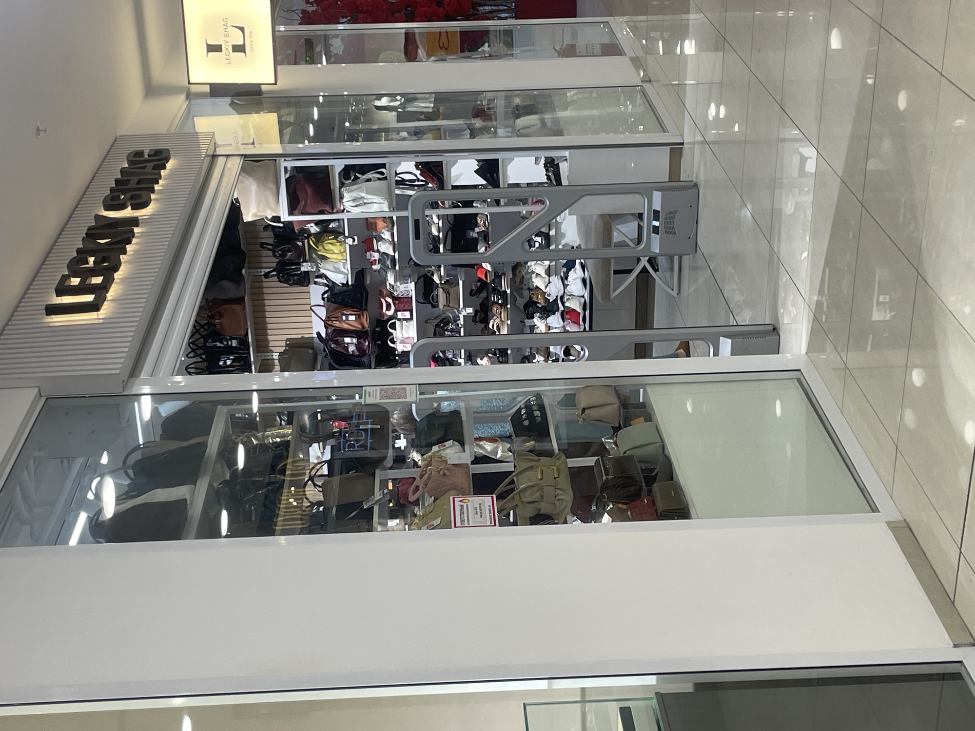 The image shows the exterior of a retail store named "LECKY SHAG," located in Tashkent, Uzbekistan. The store specializes in bags and shoes, with displays visible through a glass facade. The store's signage is illuminated, featuring the store's name in bold lettering. Inside, shelves are stocked with a variety of handbags, backpacks, and shoes. A security gate is positioned at the entrance of the store. The store appears to be situated within a shopping mall or a similar commercial complex, as indicated by the polished tiled floor and the reflective surfaces. No individuals are prominently featured or engaged in any discernible actions within the frame.