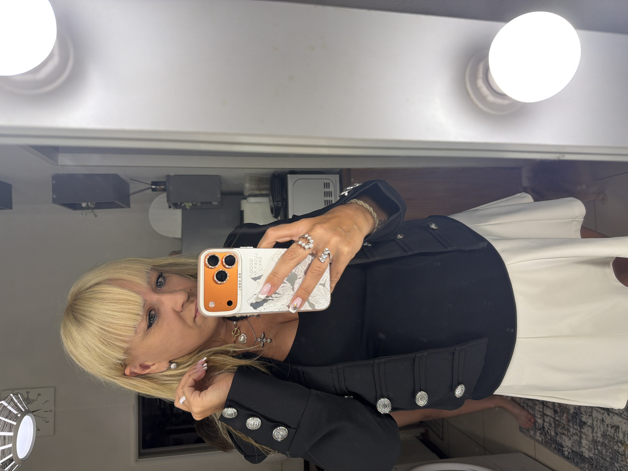A woman with blonde hair is taking a selfie in front of a mirror. The mirror has two light bulbs above it. The woman is wearing a black top and a white skirt. She is holding an orange iPhone with a decorative case. Her nails are manicured. She has jewelry on her fingers, wrist, and neck. The background shows a room with other objects visible in the mirror, including a clock, gray boxes, and a white appliance. There's no information in the image to indicate it was taken in Chaikovsky, Russia.