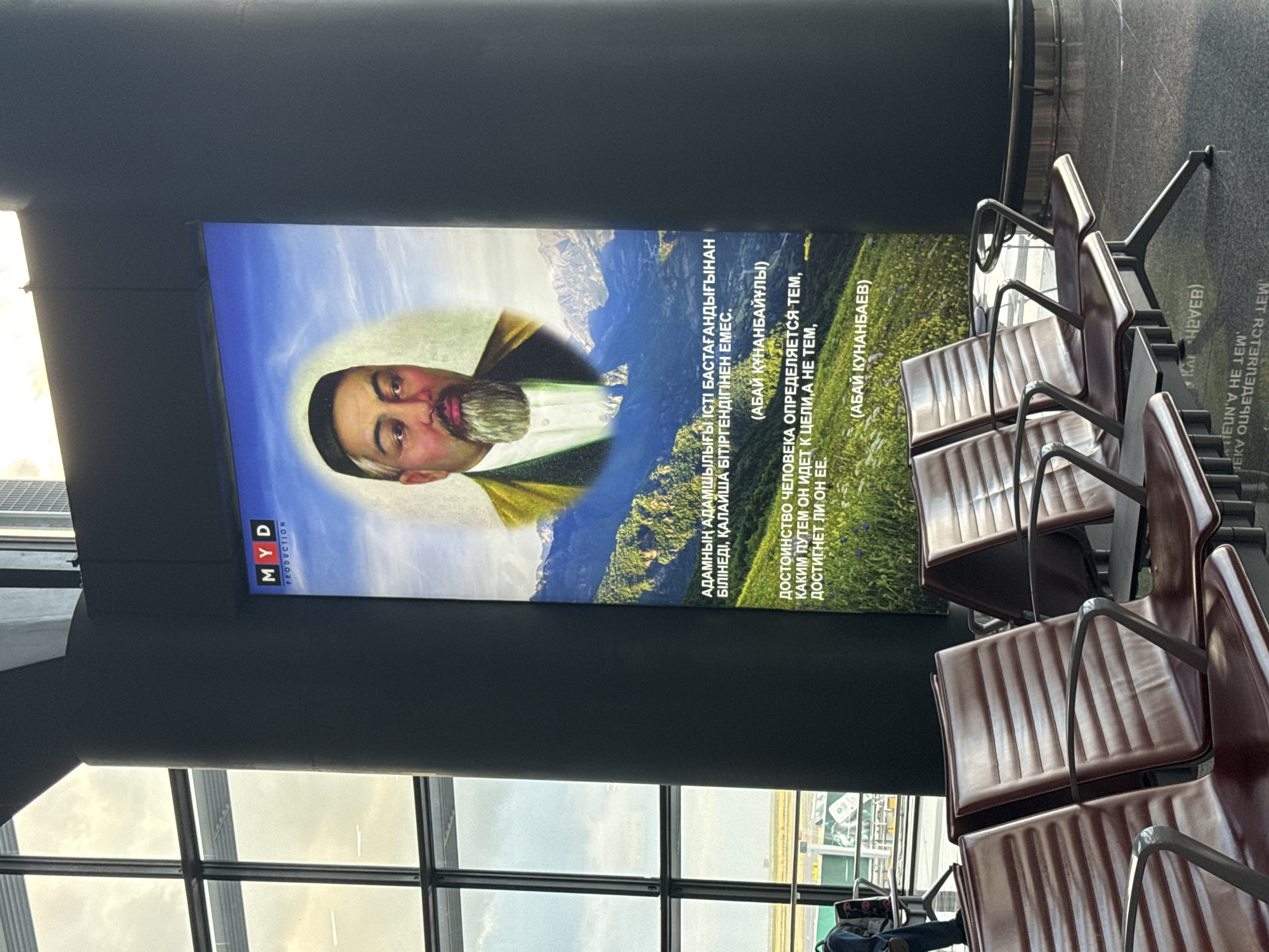 The image shows a large digital billboard in what appears to be an airport or modern public building. The billboard features a portrait of a man with a beard and a traditional cap, set against a backdrop of a mountainous landscape. Text in a foreign language, likely Kazakh or Russian, is displayed below the portrait. The billboard is flanked by large windows revealing an overcast sky and the exterior of the building, which includes a distant cityscape. In the foreground, rows of dark brown, modern-style seating are visible, arranged in a waiting area. The overall impression is one of a clean, contemporary space featuring cultural or inspirational content.