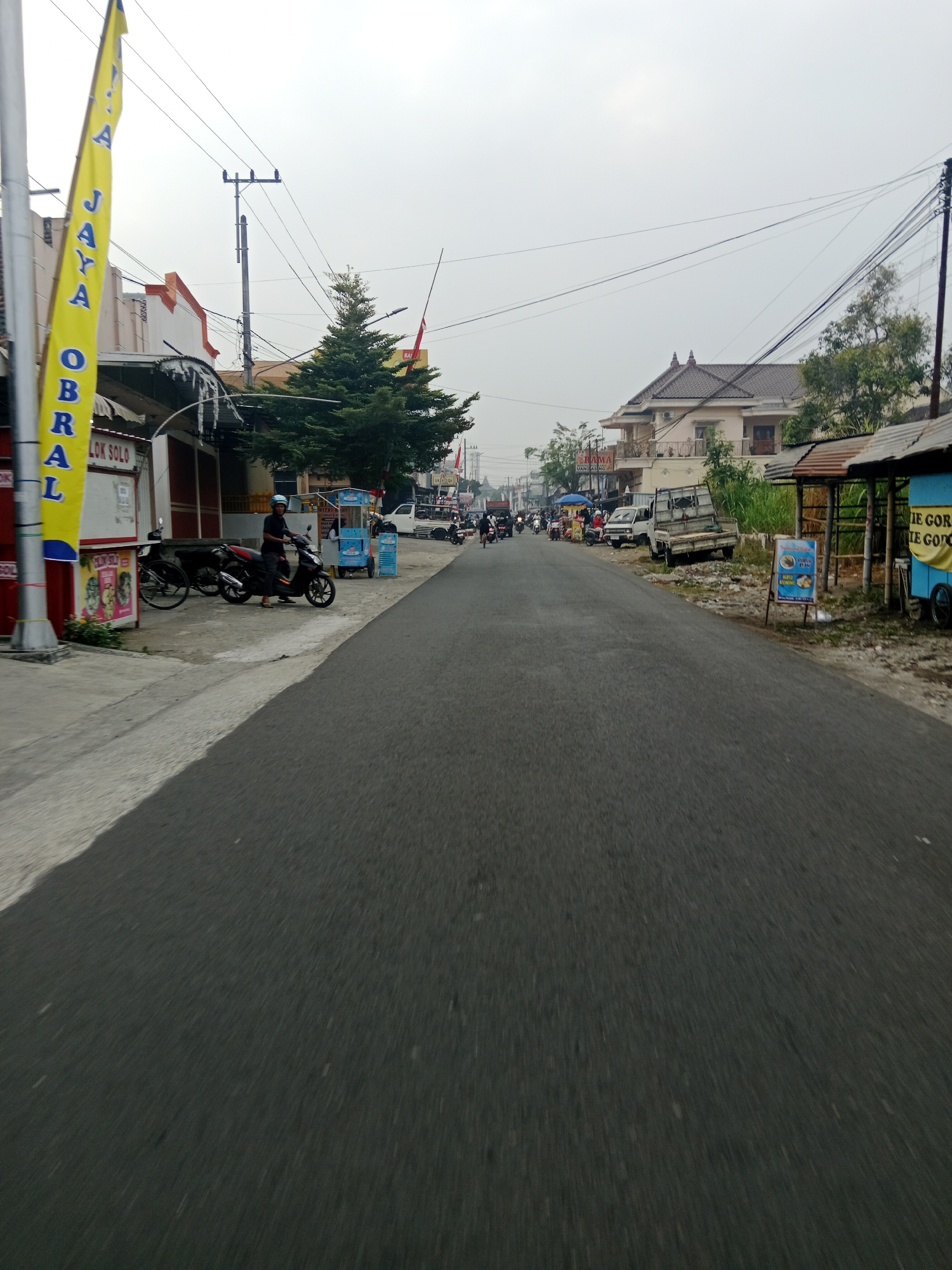 The image depicts a street scene in Srikaton, Indonesia. The viewpoint is from a moving vehicle, as indicated by the motion blur on the road. On the left side of the road, a yellow banner with blue lettering, partially visible, reads "JAYA OBRA LOK SOLO". Several buildings line the left side, with a motorbike parked in front of one. A person in a blue helmet is seated on another motorbike. Further down the street on the left, there is a small food stall with a blue and yellow cart.
The road itself is asphalt and appears to be a two-lane street. Traffic is visible in the distance, including motorbikes and a small white truck. On the right side of the road, more buildings and makeshift stalls are present, with one stall displaying a sign with blue and yellow colors. A larger building with a brown tiled roof and balconies stands further down on the right. Overhead, power lines crisscross the sky. The sky is overcast, suggesting a cloudy or hazy day. The overall environment appears to be a mix of commercial establishments and residential buildings along a local road.