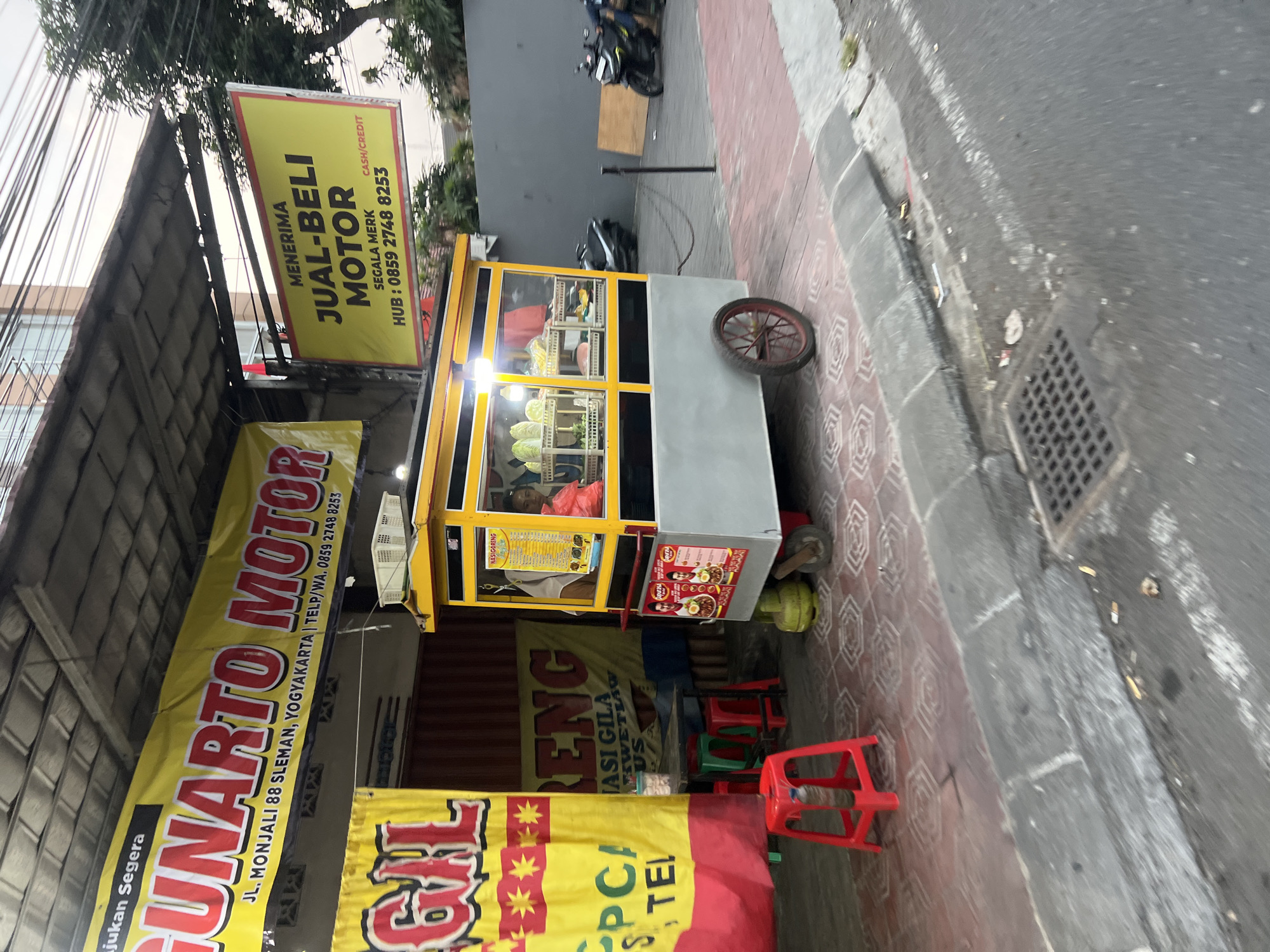 The image depicts a street scene in Sinduadi, Indonesia. In the foreground, a red plastic stool is positioned on a patterned tiled sidewalk. To the right of the stool, a yellow and gray food cart is visible, equipped with a glass display case containing fresh produce and a menu board above. A person can be seen inside the cart, attending to customers. The cart is adjacent to a red and yellow banner that advertises "GUNARTO MOTOR" with an address and phone number. Further to the right, another yellow banner displays "MENERIMA JUAL-BELI MOTOR" (Accepting Motorbike Sales and Purchases) with contact information. Several motorcycles are parked along the street, suggesting a commercial area. The environment includes a gray concrete wall in the background, overhead wires, and trees. The overall impression is a busy commercial street with food vendors and motorbike services.