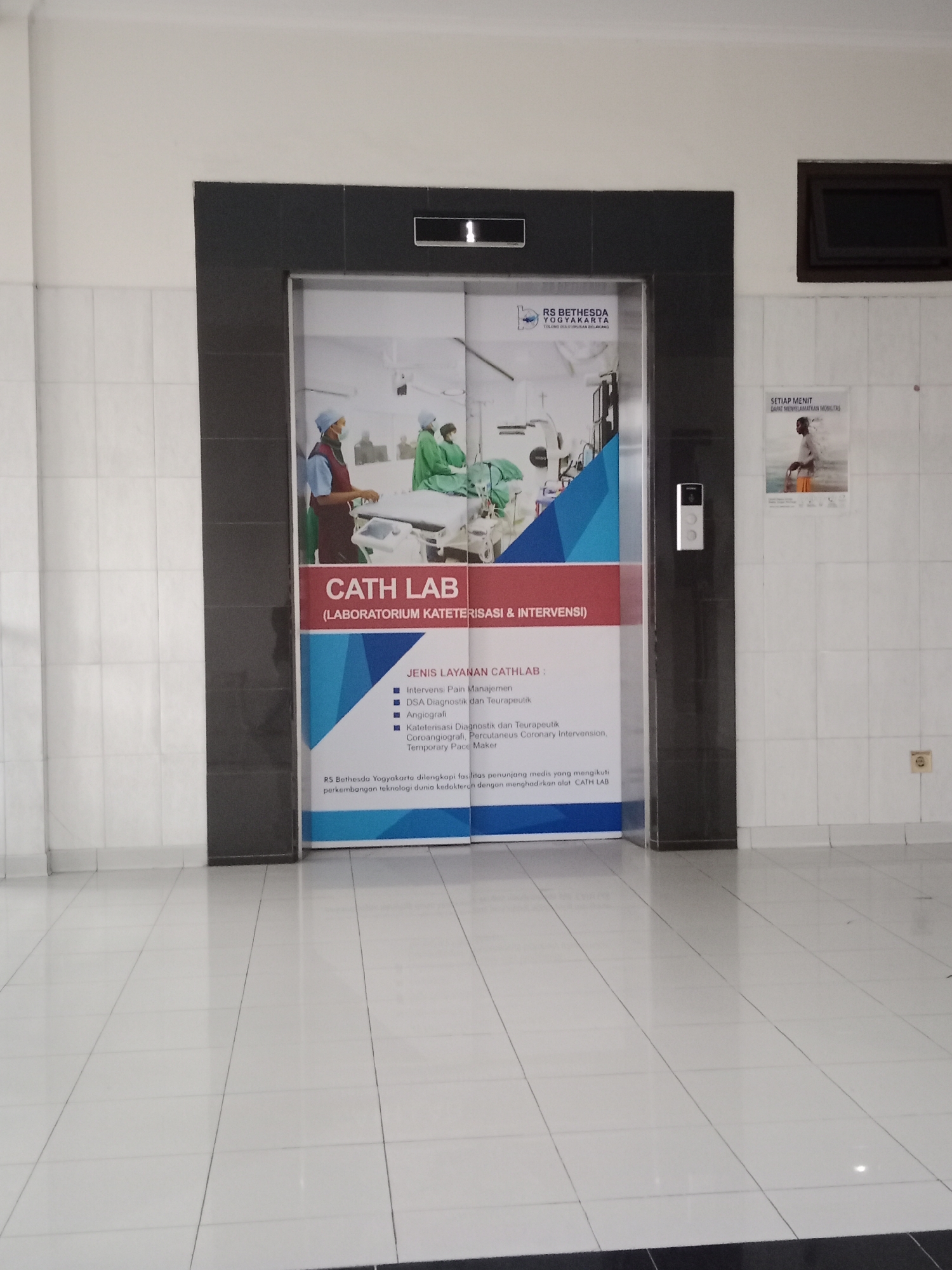 An indoor, ground-level hallway features a modern elevator with a large graphic panel on its doors. The graphic displays an advertisement for the "CATH LAB (LABORATORIUM KATETERISASI & INTERVENSI)" at RS Bethesda Yogyakarta. The image shows medical professionals in scrubs tending to a patient on a medical bed within a sterile operating room setting. The advertisement lists the services offered, including "Intervensi Pain Managemen", "DSA Diagnostik dan Teraupeutik", and "Angiografi," among others. To the right of the elevator, on a tiled wall, are two posters: one with the text "SETIAP MENIT DAPAT MENYELAMATKAN MOBILITAS" and an image of a man with a cane, and another smaller poster featuring the RS Bethesda Yogyakarta logo and an Indonesian phrase. A standard electrical outlet is visible on the wall to the right of the posters. The floor is tiled with large, reflective white tiles, creating a sense of spaciousness. The lighting appears to be artificial and even. The overall environment suggests a clean, professional medical facility.