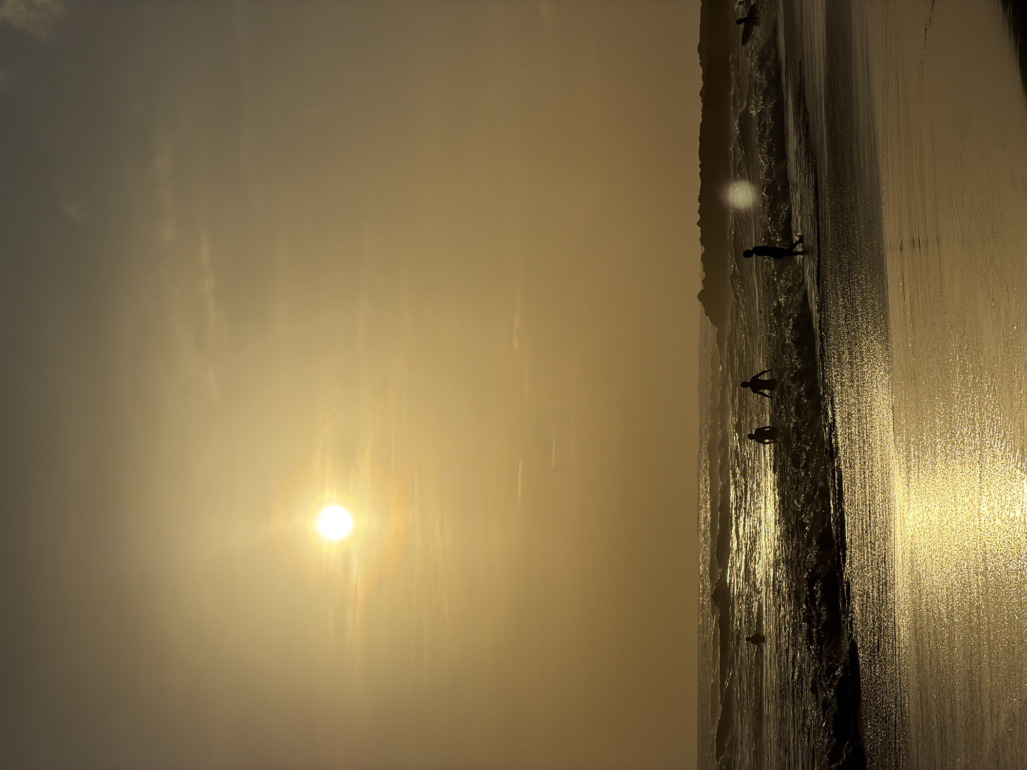 The image depicts a beach scene at sunset. The sky is a hazy golden color, with the sun prominently visible. The beach is sandy and wet, reflecting the sunlight. Several people are silhouetted along the shoreline, some appearing to be walking or playing in the water. The location is likely in North Kuta, Indonesia, based on the landscape. Waves are visible along the shore.