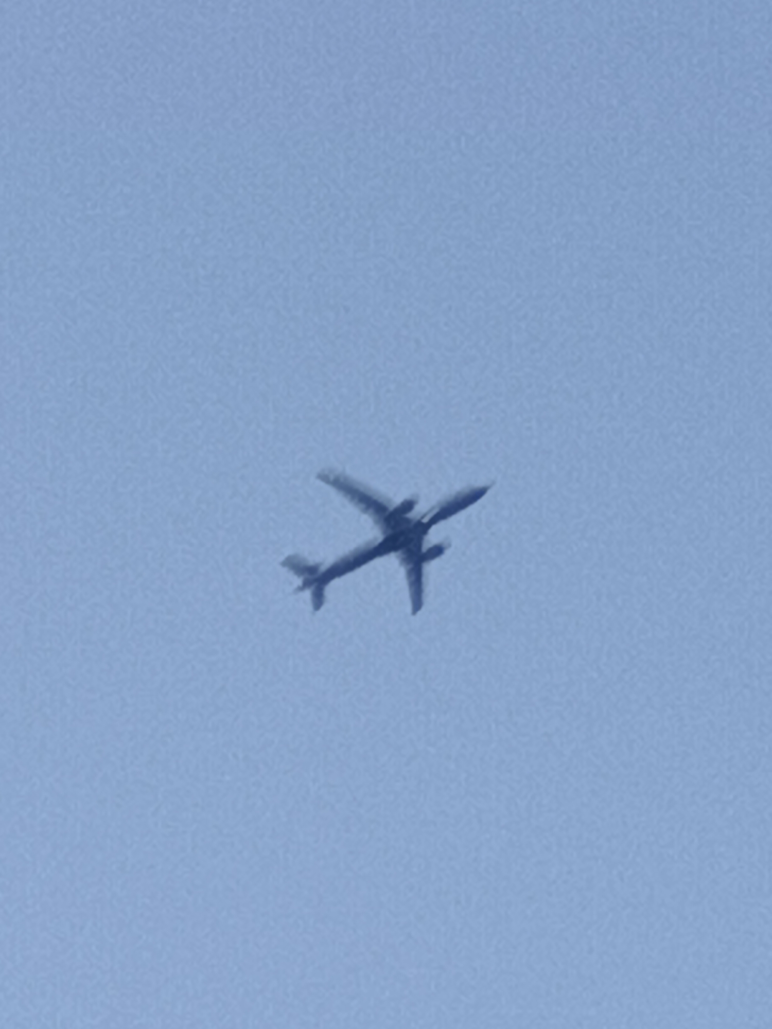 A dark silhouette of a jet airplane is captured in flight against a pale blue sky. The airplane is positioned in the center of the frame and appears to be ascending or banking slightly. Its wings are extended, and the tail section is clearly visible. The overall image is somewhat grainy, suggesting it might be a photograph taken from a distance or with low light conditions. There are no discernible details of any specific location in Prague, Czechia, as the image focuses solely on the airplane and the sky.