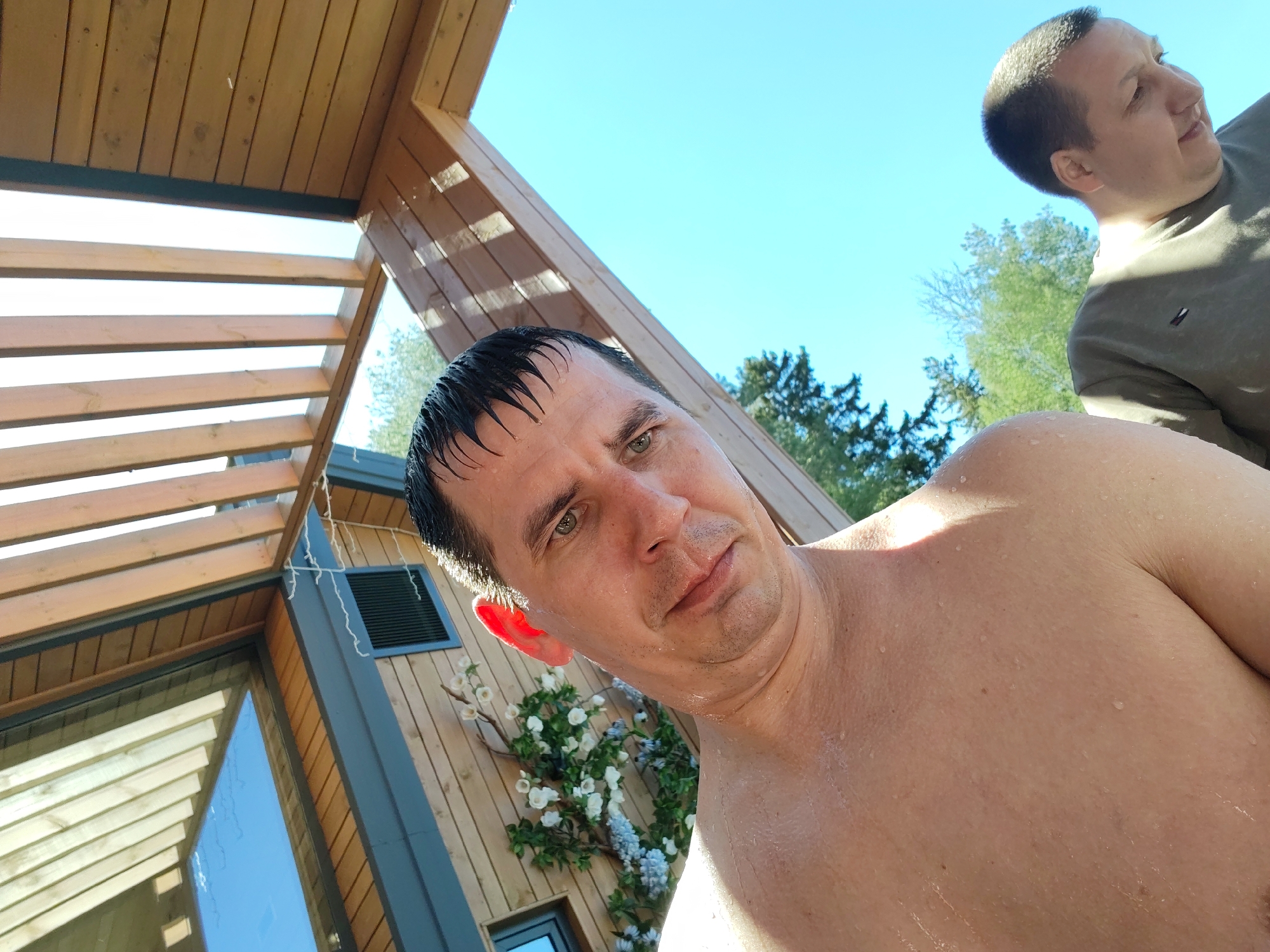A medium-angle, sunlit outdoor shot features a fair-skinned man in the foreground with wet, dark hair slicked back. He has a slight frown and is shirtless, with visible water droplets on his skin. Behind him to the upper right, another man with a similar complexion and short hair is partially visible, wearing a brown shirt.
To the left, there is a wooden structure with a slatted roof and glass windows. The side of a wooden building is visible, adorned with a floral decoration. The sky is clear and blue.
While the prompt asked for the location to be Izhevsk, Russia, there is nothing in the image itself to conclusively verify this. The scene appears to be a private outdoor setting, possibly a backyard or patio with a sauna or bathing facility.