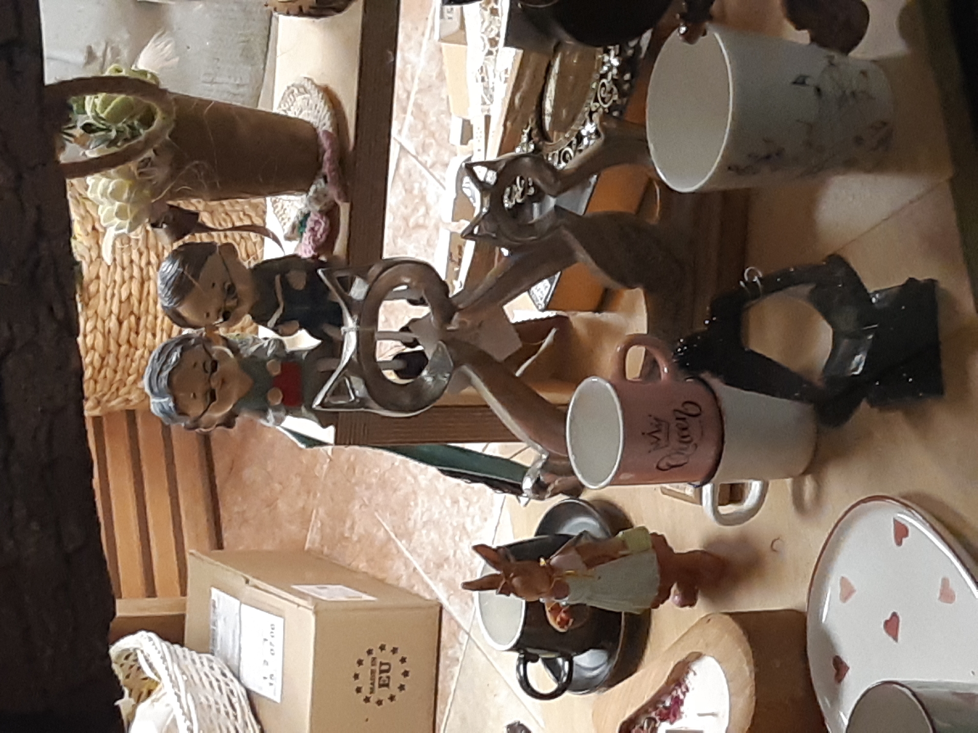 The image shows a collection of assorted objects on a floor. These include a pair of porcelain figurines, a decorative wooden sculpture, several mugs (one pink with the text "My Queen"), a decorative bunny figurine on a mug, a plate with red hearts, a small basket with artificial flowers, and a cardboard box. The cardboard box has "EU" printed on it, along with an unreadable date or code: "15 1 07 00". The items are placed in what appears to be a corner or a niche within a room. Specific information indicating that the photo was taken in Plovdiv, Bulgaria is not evident in the image.