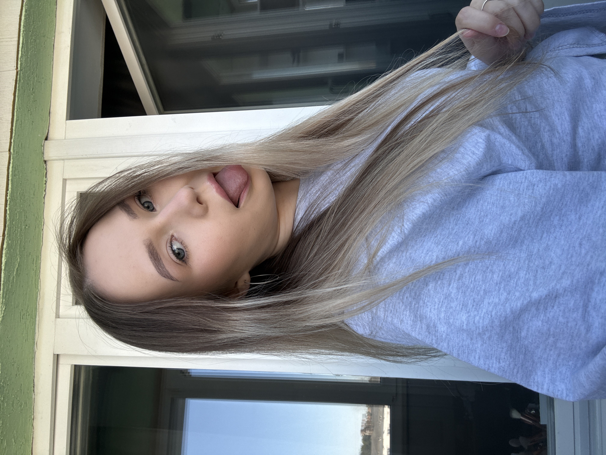 A fair-skinned woman with long blonde hair is sticking her tongue out at the camera. She is wearing a gray sweatshirt. The photo appears to be taken indoors near a window with a view of what is most likely a city environment outside. While the photo provides no specific geographic markers, it is said to have been taken in Chaikovsky, Russia. This additional information does not alter the description of the scene as presented in the image.