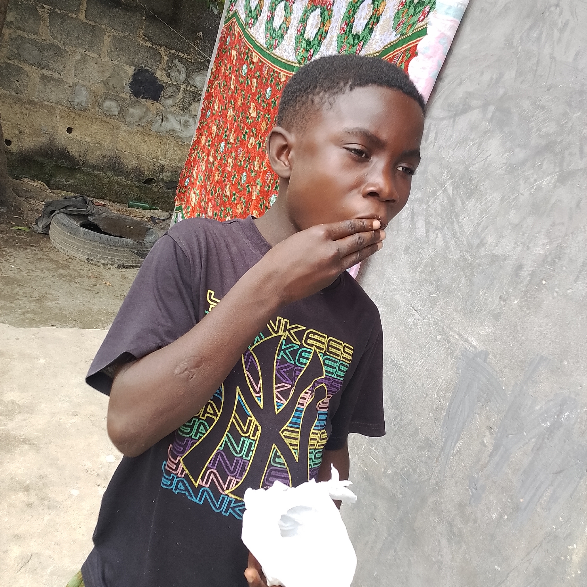 A young Black male, appearing to be around 10-12 years old, stands outdoors in what is likely a residential area in Ado, Nigeria. He is wearing a black t-shirt with a colorful graphic design. His right hand is raised to his mouth, and he appears to be eating something white, possibly a piece of food or candy. He is looking to his right, with his gaze slightly downward.
The background consists of a textured grey wall on the right, possibly concrete, with some scuff marks and faint grey brush strokes. To the left of the wall, a colorful tapestry or fabric with a red and green patterned design is draped. Further to the left, a rough, greyish-brown stone wall is visible, with some darker patches and small holes. On the ground, a discarded tire is visible, with some dark fabric placed on top of it. The ground itself is a mixture of dirt and concrete. There is a green bottle lying on the ground near the tire. The overall environment suggests an outdoor, possibly semi-urban or rural setting.