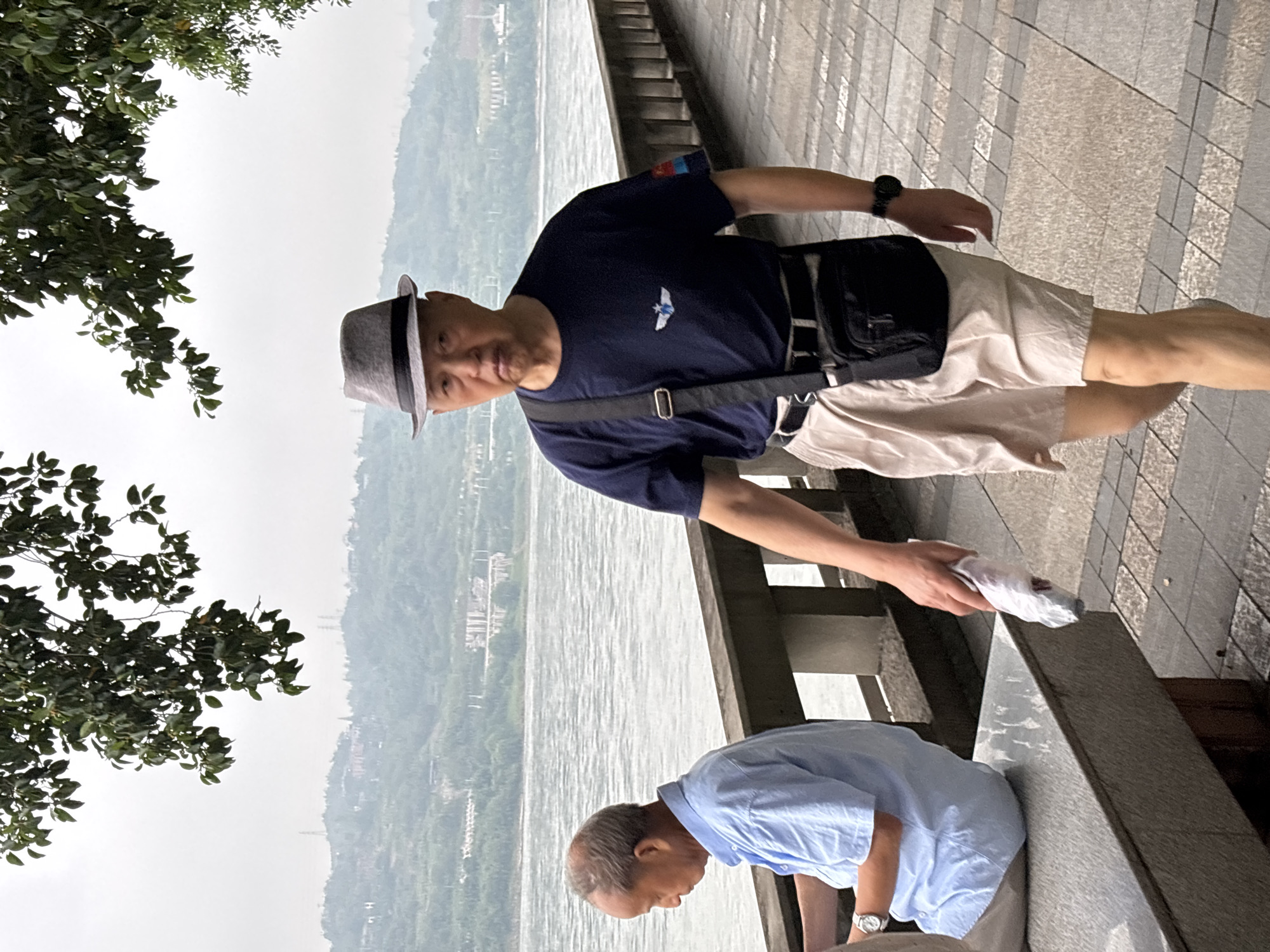 In the city of Jiangjin, China, an elderly man wearing a gray fedora, a dark blue t-shirt with a small emblem, and light-colored shorts walks along a paved promenade. He has a black crossbody bag and a watch on his left wrist. He is holding a white plastic bag in his right hand. Behind him, another elderly man in a light blue short-sleeved shirt and khaki pants sits on a bench, looking down. The promenade runs alongside a wide, calm river. In the background, a hilly landscape with scattered buildings and vegetation is visible under an overcast sky. Trees with green leaves hang over the scene from above.