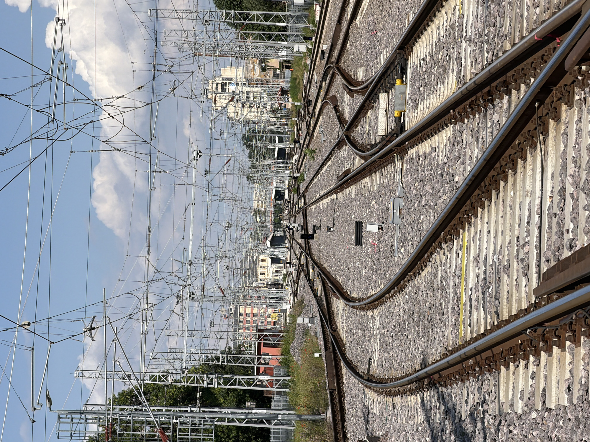 The image shows a network of railway tracks converging in Plovdiv, Bulgaria. The tracks are laid on a bed of gravel and wooden sleepers. Overhead power lines supported by metal pylons stretch along the tracks, creating a complex web of wires against a backdrop of a blue sky with white clouds. Buildings, some residential, are visible along the line, their design representative of Plovdiv architecture. The scene suggests a railway junction, likely a point of transition or interchange within the city's transportation network. No people are visible in the frame.