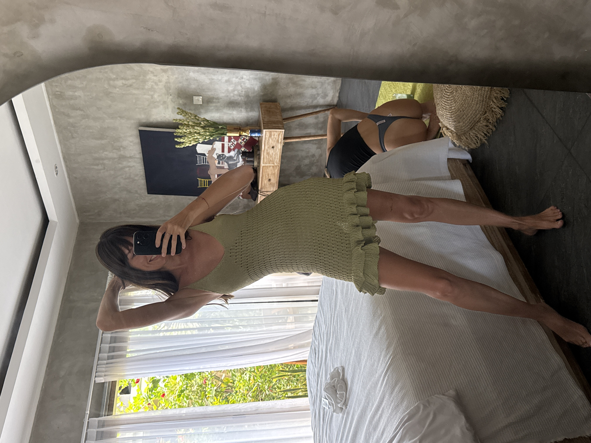 A vertical mirror selfie, taken indoors in North Kuta, Indonesia, captures two individuals. The primary subject, a woman with dark hair and bangs, is seen reflected in the mirror. She wears a light green, possibly crocheted or knit, mini-dress with ruffles along the side. She holds a dark smartphone to her face, partially obscuring it, but her eyes and a slight smile are visible. Her other arm is bent, resting behind her head, and she stands with one leg extended slightly.
In the background reflection, behind the main subject, another person is seen from behind, bent over on a bed. This individual is wearing dark underwear (a thong) and a dark top, appearing to be stretching or posing. A straw-woven bag rests beside them on the bed.
The setting appears to be a modern bedroom or studio. The room features light gray, textured walls and dark gray tiled floors. Behind the bent-over person, a light wooden side table with drawers holds a vase with tall, light-colored flowers or foliage. A dark wall hanging with abstract or cartoon-like designs (including what looks like a cat) is visible above the table.
Natural light floods the room from a large window covered by sheer white curtains. Lush green foliage and hints of red flowers are visible outside, indicating a sunny daytime. A white, waffle-textured blanket covers the bed, and a white scrunchie or hair tie lies on it near the main subject's feet. Text, possibly "IPHONE," is partially visible on the main subject's phone case. Small, indistinct text is also visible on the waistband of the underwear worn by the second person.