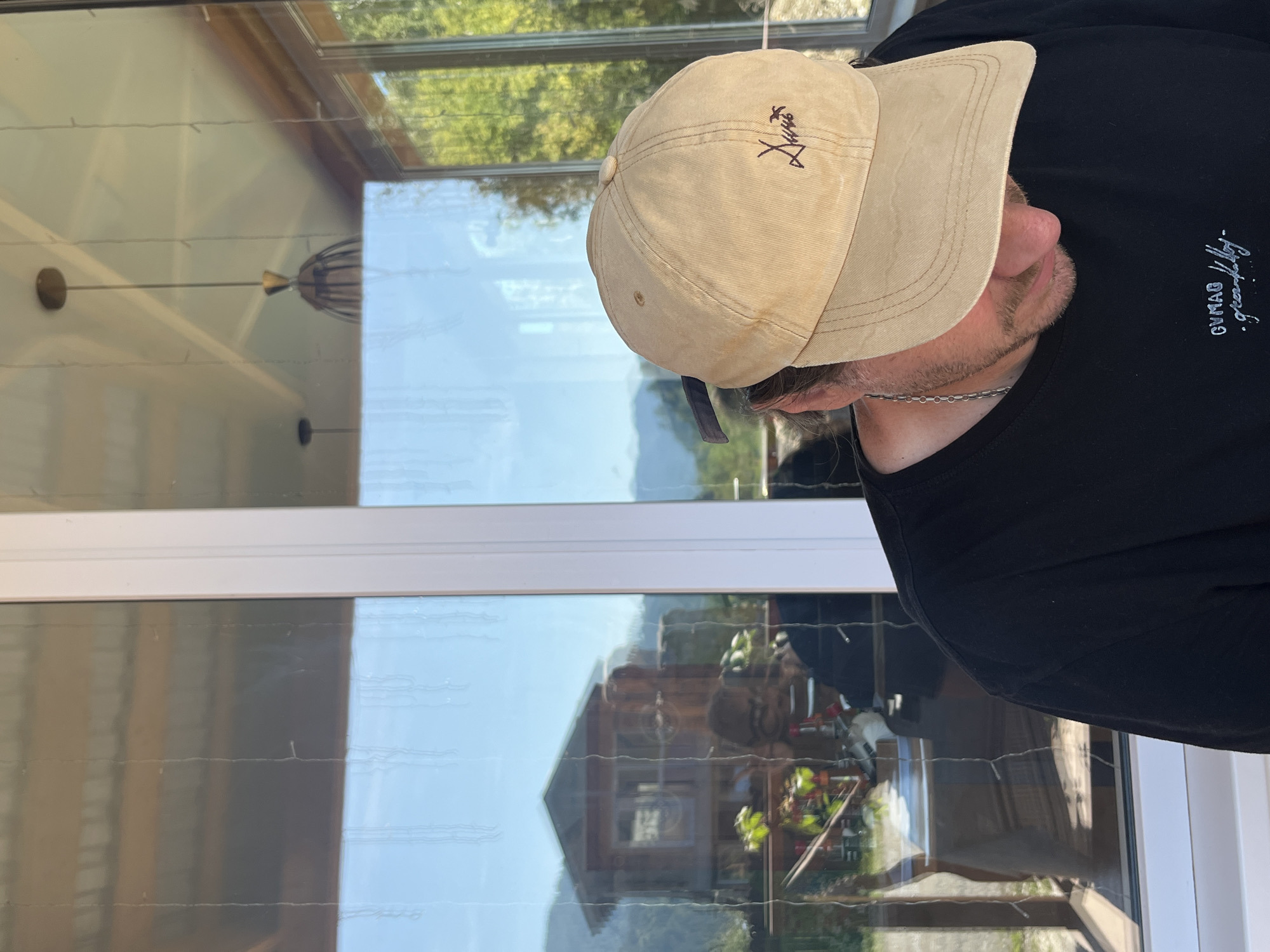 The image shows a person wearing a black t-shirt and a tan baseball cap, looking down. The person is in an indoor setting with large windows reflecting the outside environment. The reflection shows a wooden structure that appears to be a house on stilts, surrounded by trees and mountains, suggesting a rural or mountainous location. The text on the t-shirt and the cap are partially visible. The overall lighting suggests it is daytime. While the image does not explicitly state the location, it is implied to be Ust-Muny, Russia.
