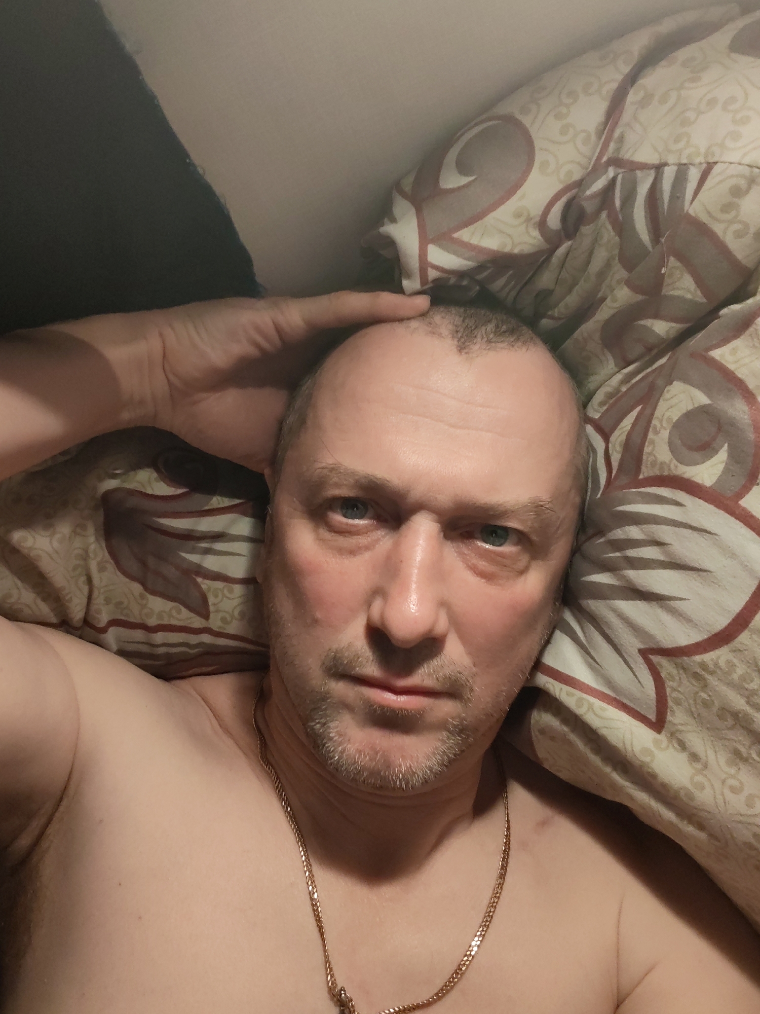 A middle-aged Caucasian man with thinning hair and a stubble beard is lying on a bed covered in a patterned beige and brown comforter. He is shirtless and wearing a gold chain necklace. His right hand is touching his forehead. The background is out of focus, but a dark object, possibly a pillow, is visible in the upper left corner. The location in Topornya, Russia cannot be determined from the image.