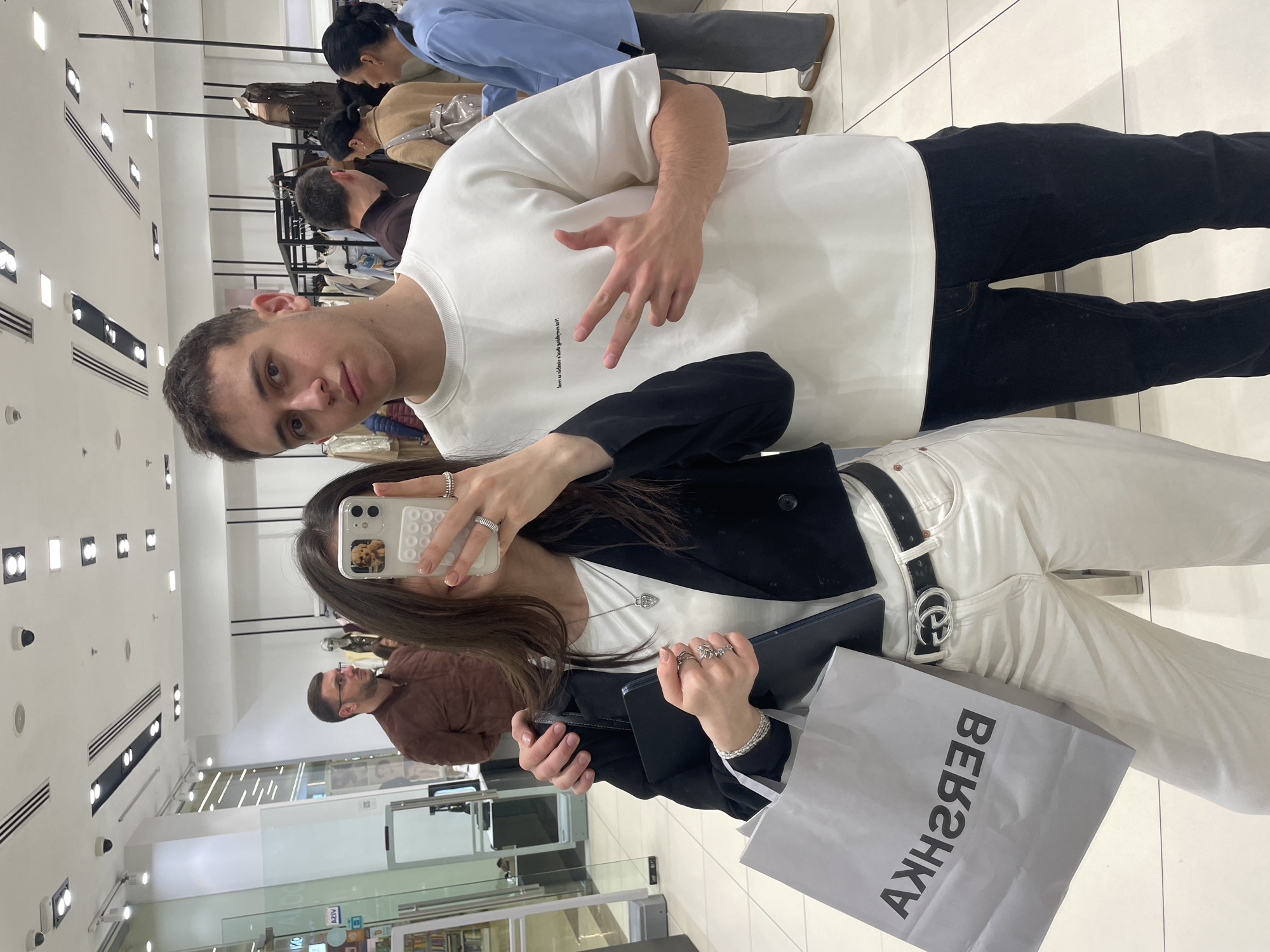 Two young adults are posing for a selfie in a clothing store. The man is on the left, wearing a white t-shirt and black pants, holding up two fingers. The woman is on the right, holding a cell phone with a dog image on the screen, a tablet, and a Bershka shopping bag. She's wearing white pants, a white shirt, and a black blazer. The store's interior is visible in the background, with clothing racks and other customers. The store appears to be located in Yerevan, Armenia.