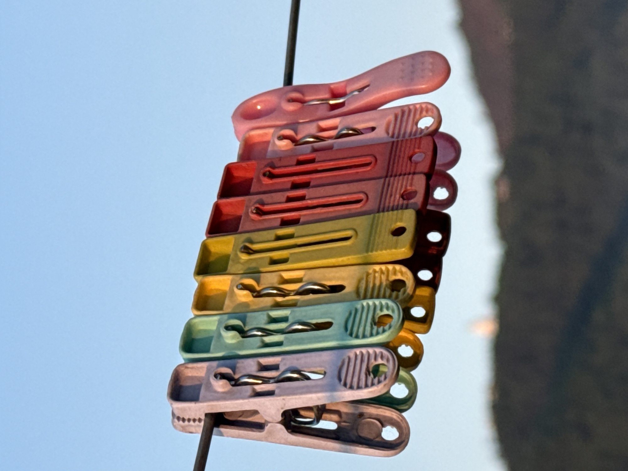 A close-up shot shows a stack of multi-colored plastic clothespins hanging on a wire line. The colors of the clothespins range from pink at the top, to red, yellow, green, and light purple at the bottom. A thin wire runs vertically through the clothespins. The background is a clear, light blue sky on the left and a blurred view of a dark green mountain or hill on the right. The photo was likely taken in Pokhara, Nepal.