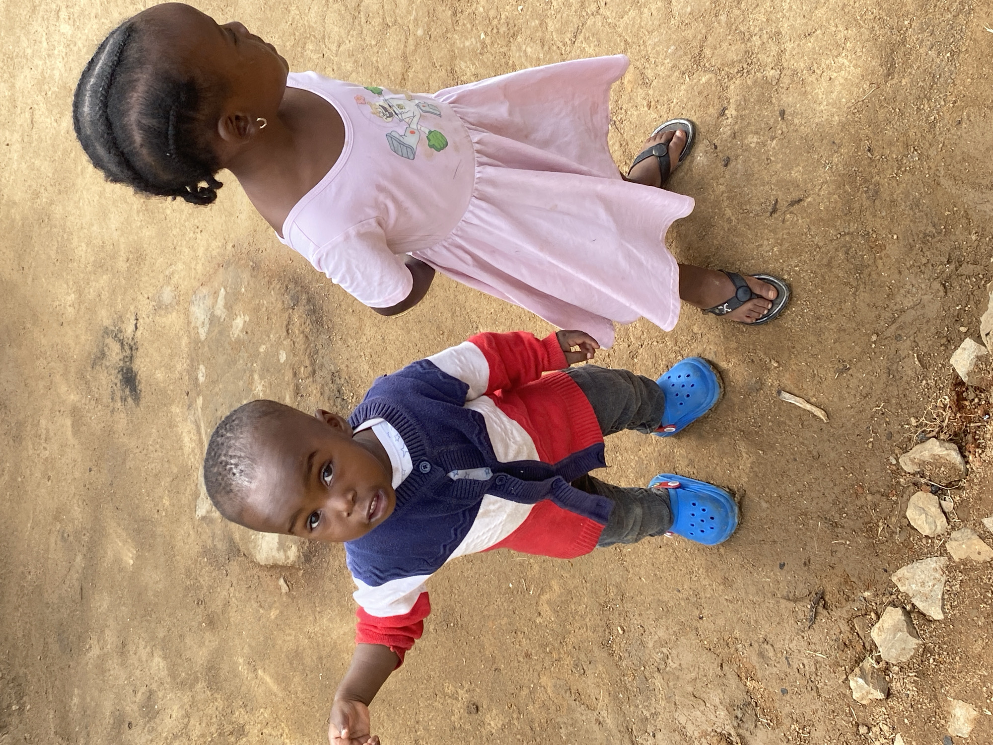 Two young children are standing on a dirt ground. The girl is above and to the right of the boy. She is wearing a pink dress and sandals. The boy is wearing a red, white, and blue sweater, gray pants, and blue Crocs. The ground is dirt and has rocks on the right. The location is Jos, Nigeria.