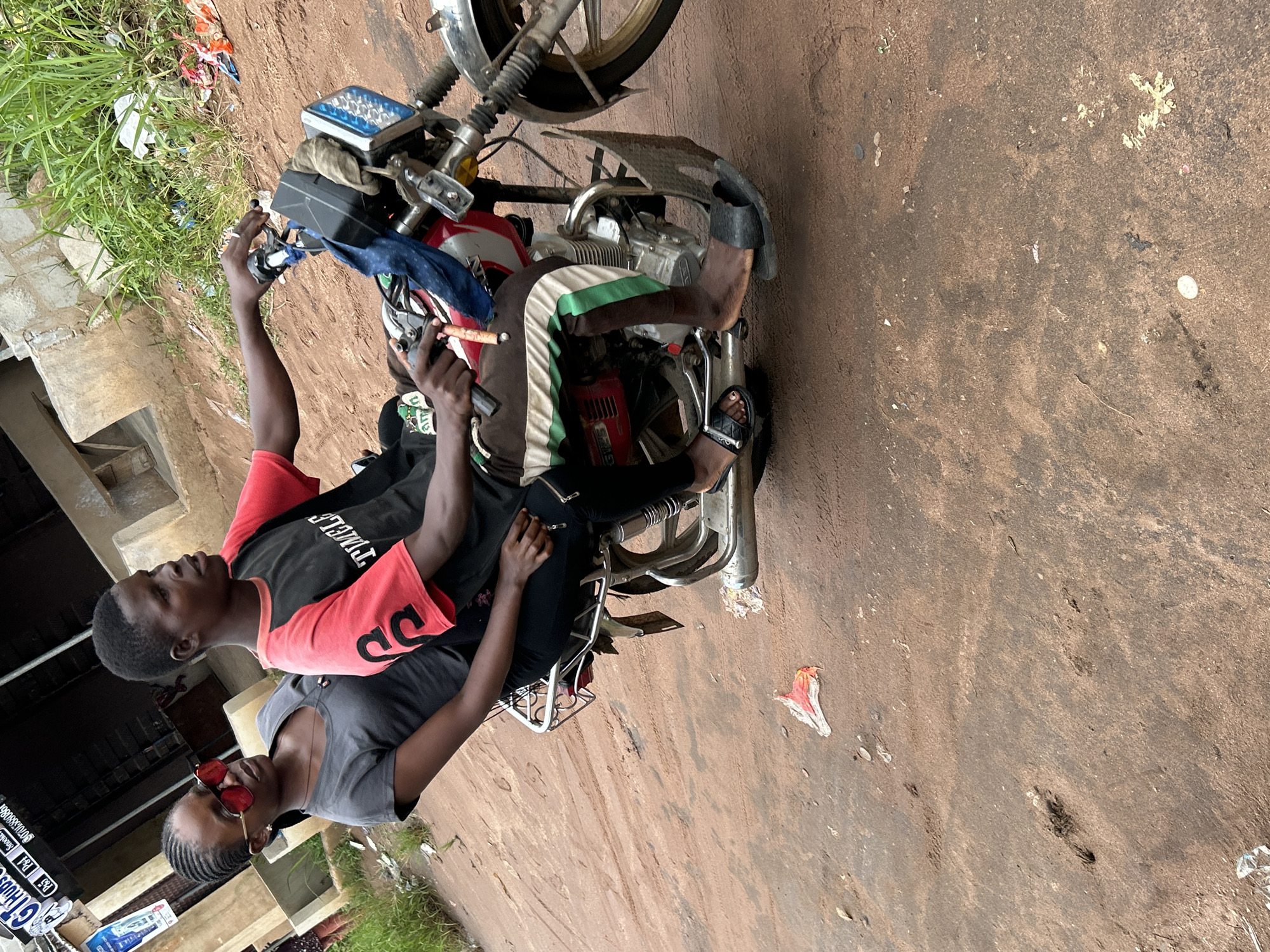 Two individuals are on a red and silver motorcycle, with the driver in front and a passenger behind. The driver, a young male with dark skin, wears a black t-shirt with red sleeves and brown pants with green and white stripes. The passenger, a young female with dark skin and braided hair, wears a gray t-shirt and red-tinted sunglasses. Both are looking forward. The motorcycle is stopped on a dirt road, which appears to be in a semi-urban or rural setting. The background shows a building with concrete walls and some overgrown green grass and debris. There are tire tracks visible in the dirt road, suggesting recent vehicle movement. The scene is likely in Ihiagwa, Nigeria, given the context.