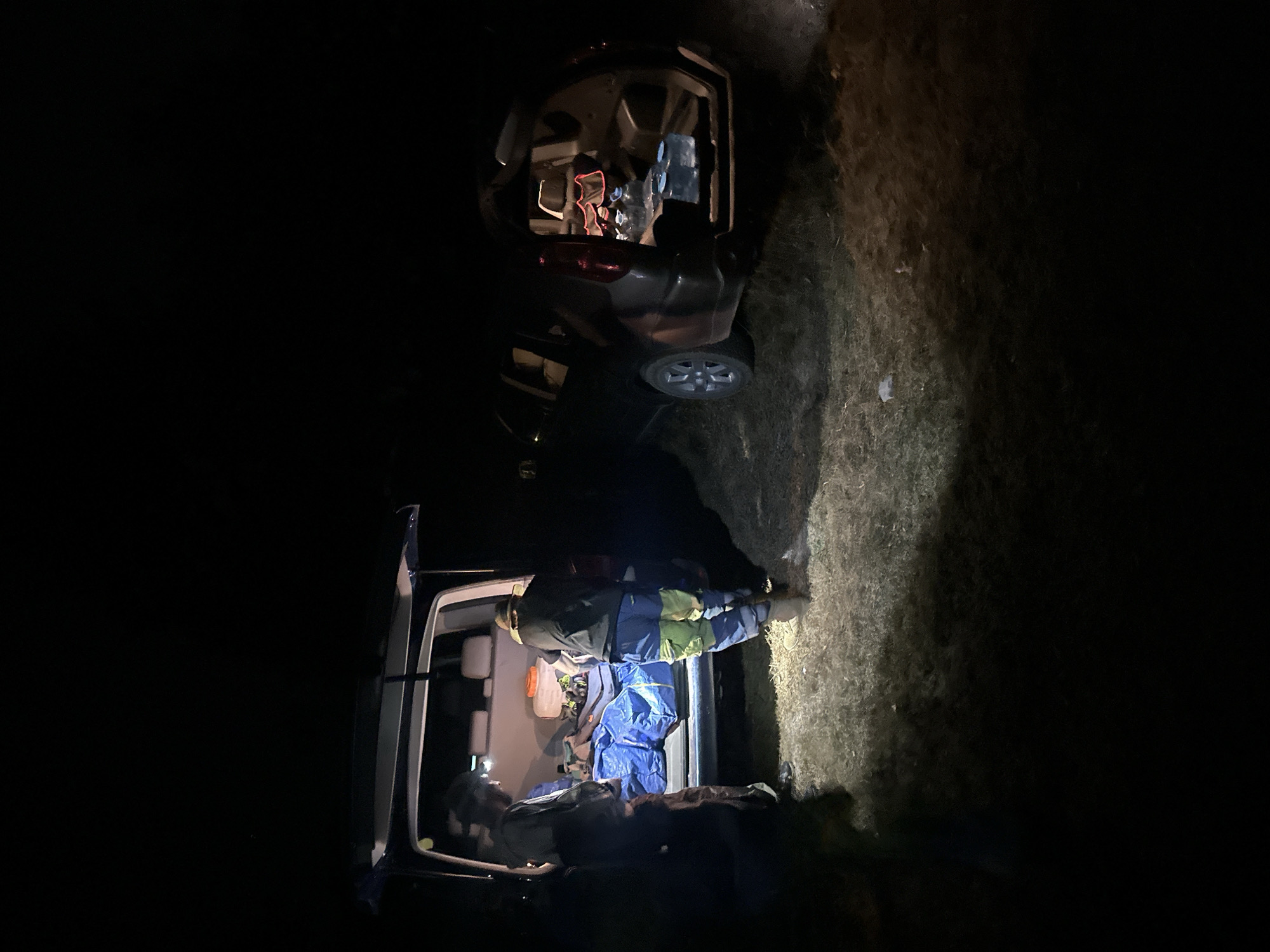 At night, two individuals are visible near the open rear of a dark-colored SUV, its tailgate lifted, revealing several large plastic water jugs within. Adjacent to the SUV, the rear doors of a blue van are also open, with a blue tarp spread across the floor and what appear to be backpacks or bags on top of it. The ground is covered in grass and dry vegetation, illuminated by artificial lighting that casts shadows. The scene suggests preparation or unloading of supplies. The location is identified as Bijele Poljane, Montenegro.