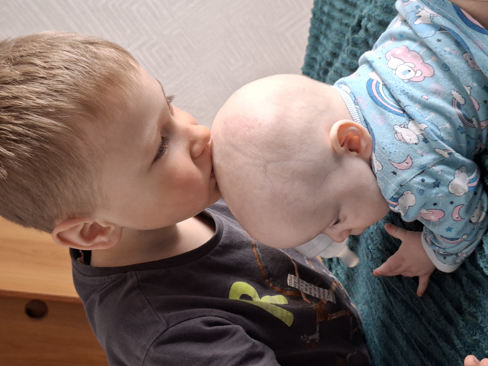 A close-up shot shows a young boy kissing a baby on the head. The older boy, positioned on the left, has short brown hair, and is wearing a dark gray t-shirt with some light green and brown graphics. The baby, positioned on the right, is wearing blue pajamas with a unicorn pattern and is lying on a teal-colored blanket. The baby’s head is bare, and only a small portion of the baby's face is visible. The setting appears to be indoors. A portion of a wooden table or cabinet is seen in the lower-left corner of the frame. The background includes a white wall with a faint pattern. The location is Unknown, Unknown.