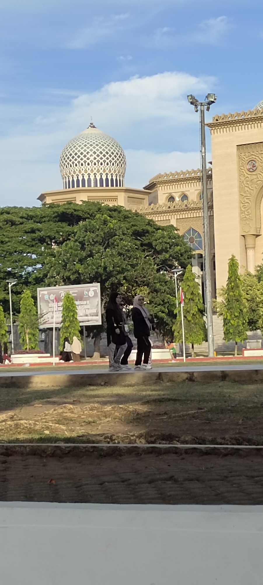 The image depicts the exterior of the Islamic Center of Lhokseumawe, a prominent landmark in the city of Lhokseumawe, Indonesia. The mosque features a large, ornate dome with a patterned facade, and it is flanked by a sandy-colored building with intricate architectural details and arched windows. In front of the mosque, a lush canopy of mature trees provides a verdant backdrop.
In the foreground, three individuals, presumably women, are walking from left to right across a paved walkway. They are all wearing Islamic attire, including hijabs. Two of them are dressed in dark clothing, while the third wears a lighter-colored hijab and a dark outfit. Their posture suggests they are casually strolling.
Behind the walking individuals, a large billboard with text and graphics is visible, indicating a public event or announcement. Several slender, conical trees are planted along the walkway. In the far background, a street lamp with multiple lights stands tall against the blue sky, which is dotted with scattered clouds. The ground in the immediate foreground appears to be a mixture of soil and sparse vegetation. The scene conveys a sense of urban tranquility, with the architectural grandeur of the mosque serving as a focal point.