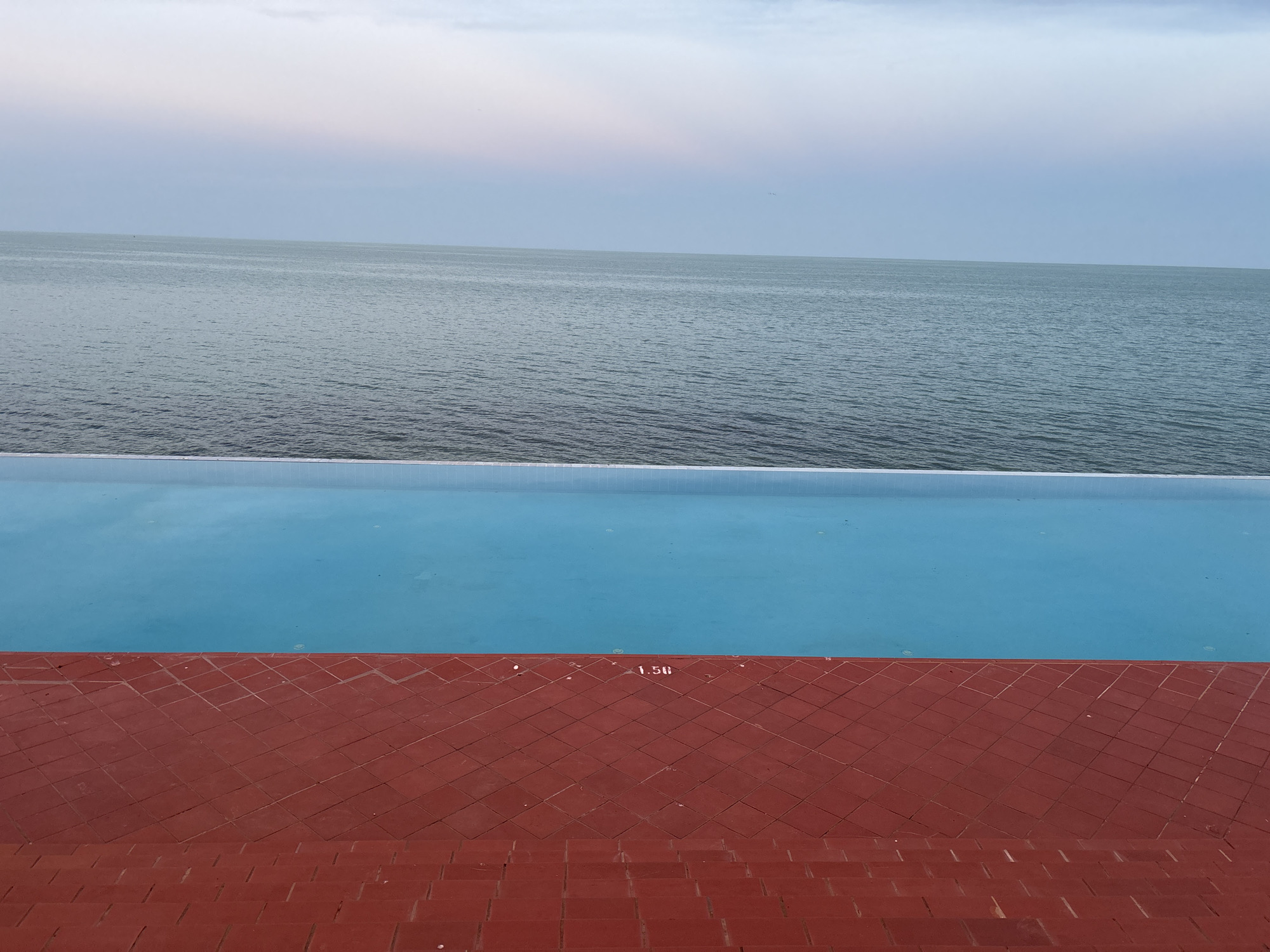 The image depicts a view of the Black Sea from an infinity pool in Varna, Bulgaria. The foreground shows a red tiled terrace leading down to the pool. The pool is filled with bright blue water, and a depth marker of "1.50" is visible on the tiling at the edge of the pool. The midground features the calm, blue-green water of the sea, stretching to the horizon under a pale, overcast sky with hints of pink and light blue. The overall scene conveys a sense of tranquility and a seaside resort atmosphere.