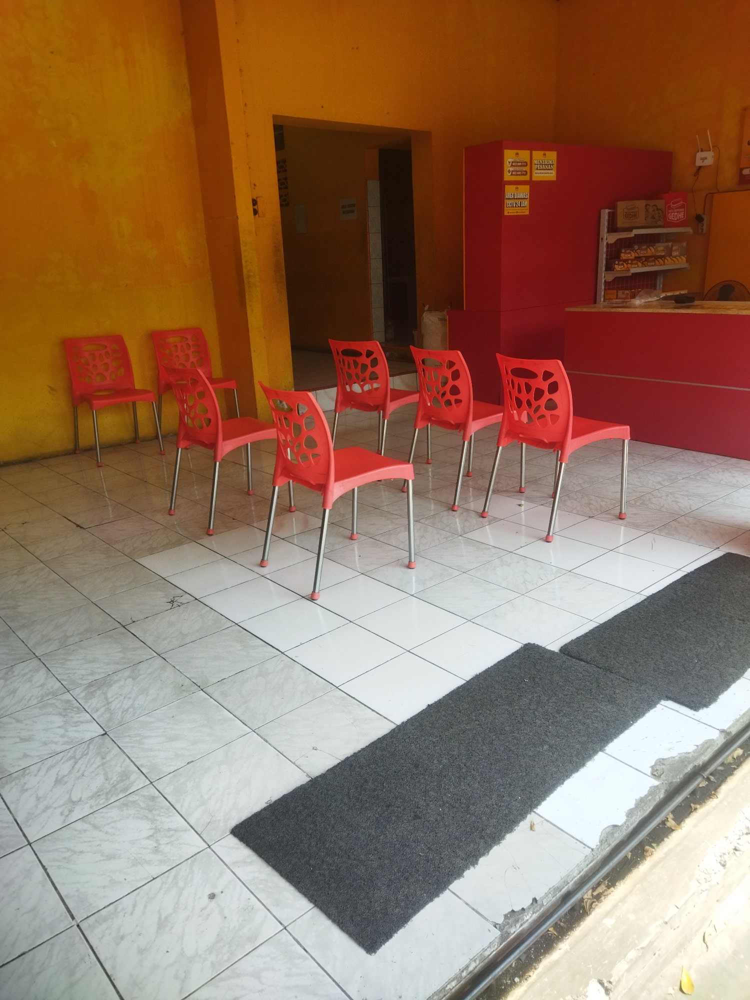 The image shows an outdoor seating area with a row of bright red plastic chairs with metal legs and circular cutouts in the backrests. The chairs are arranged on a tiled floor with a white, marble-like pattern. In the background, there is a bright yellow wall and a red structure that appears to be a counter or service area. Shelves are visible on the red structure, displaying what look like packaged food items. Signs are posted on the yellow wall and the red structure, some with text in Indonesian. Two dark grey mats are laid out on the tiled floor. The overall scene suggests a casual dining or waiting area, possibly at a food stall or small eatery. The presence of Indonesian text on the signs indicates the location is in Indonesia, and the query specified Wonogiri, Indonesia. The image captures a still moment with no people present and no apparent action.