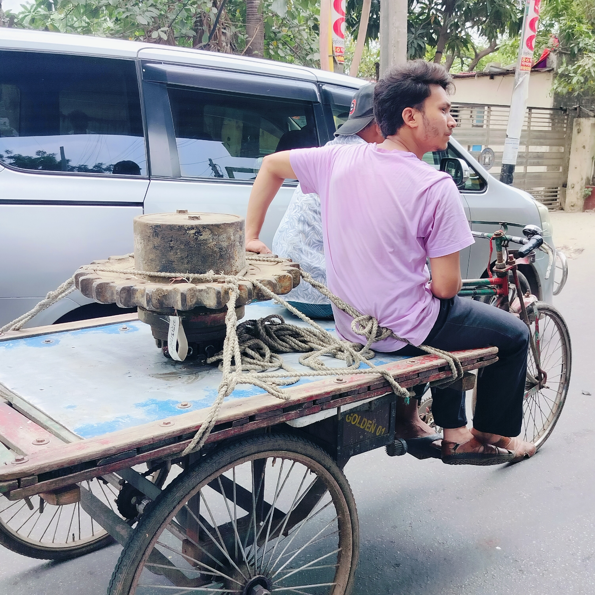 The image captures a scene in what appears to be an urban street in Unknown, Unknown. At the center of the frame is a man in a pink t-shirt and dark pants, seated on a three-wheeled cart. He is facing to the right and wears sandals. The cart is laden with a large, rusty metal gear, secured with rope. Another person, partially visible, sits behind the man. A silver van is parked nearby. The environment includes trees and buildings visible in the background.
