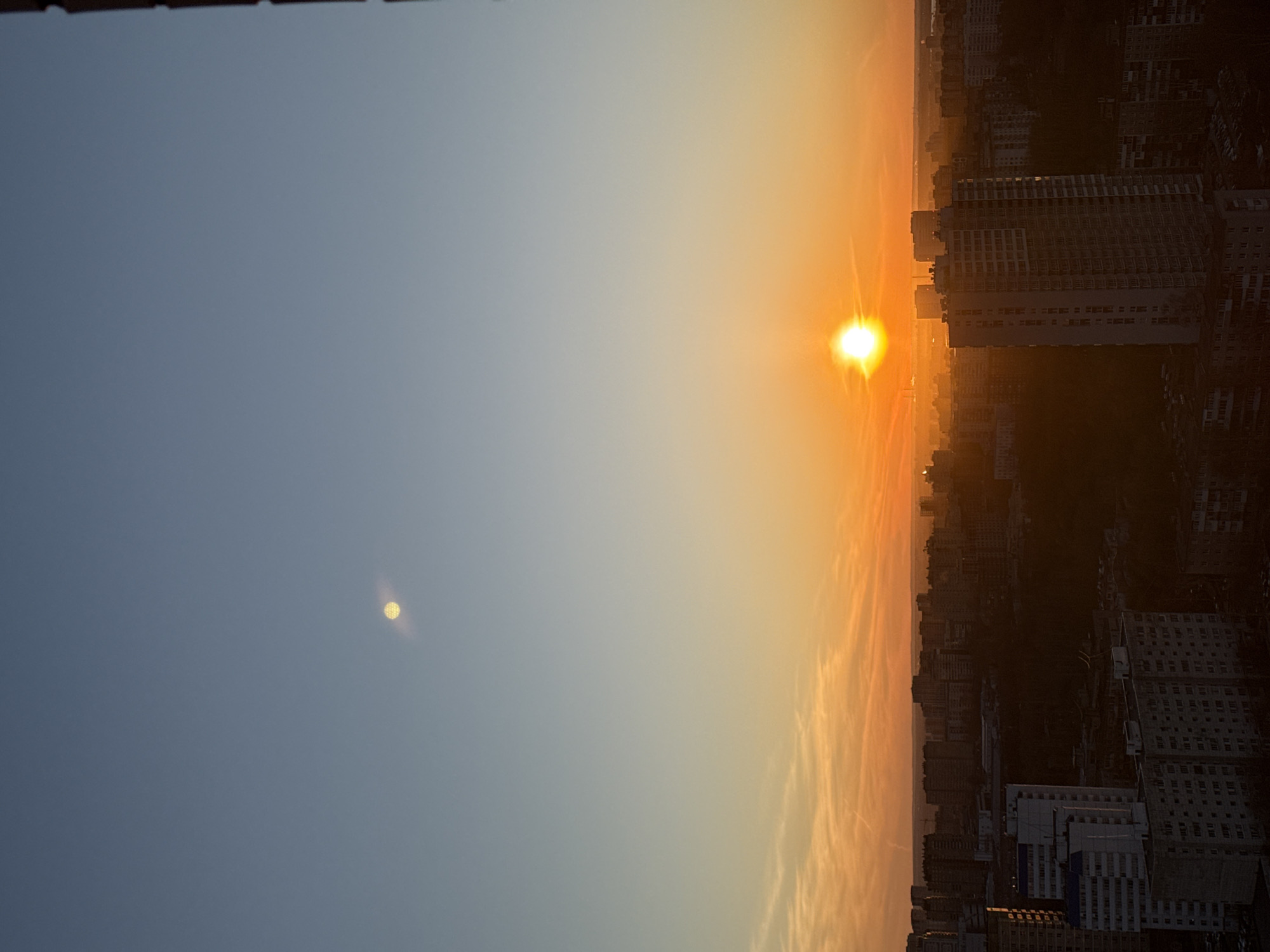 Here's a description of the image:
The image shows a sunrise over Perm, Russia. The sky transitions from a pale blue on the left to a bright orange and yellow near the horizon, where the sun is positioned. A second bright spot, possibly a reflection of the sun, is seen in the sky to the left of the sun. The cityscape of Perm is visible on the right side of the image, silhouetted against the bright light of the sunrise. Buildings of varying heights are distinguishable, with the silhouette of taller buildings are more visible in the foreground. The lighting suggests early morning, with the sun low on the horizon.