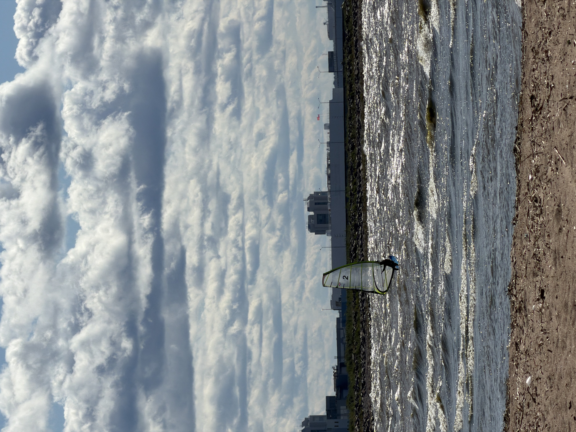The image depicts a windsurfer on the water near Kronstadt, Russia. The windsurfer is positioned on the right side of the frame, facing towards the left. They are equipped with a sail that has the number "2" visible on it, and are wearing a dark helmet and a blue top. The water is choppy with sunlight glinting off the waves. In the background, a rocky shoreline separates the water from a cityscape with buildings and streetlights. Above the city, a dramatic sky filled with large, layered clouds stretches across the frame. The clouds are predominantly white and grey, with some appearing darker, suggesting a dynamic weather system. The overall lighting indicates it is daytime, possibly late afternoon due to the angle of the sunlight reflecting on the water. The scene suggests an active recreational pursuit in an urban coastal environment.