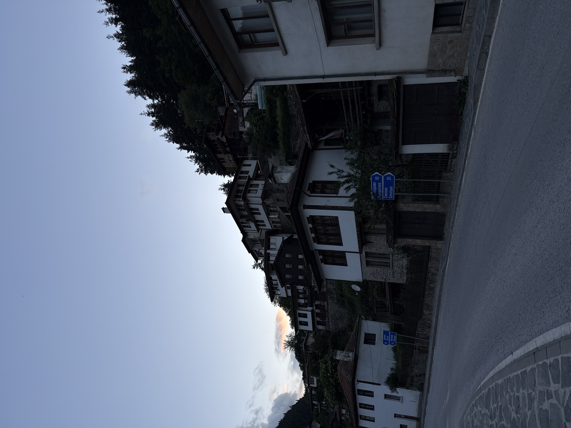 The image depicts a view of the village of Shiroka Laka, Bulgaria, with a focus on its characteristic architectural style and the hilly terrain. The scene is captured during dusk or dawn, as indicated by the soft, diffused light and the clear, pale sky.
Dominating the frame are traditional Bulgarian houses, built on steep slopes. These structures are characterized by their white-washed walls, dark wooden accents, and red-tiled roofs. Many of the houses feature multiple stories, with balconies and upper levels jutting out, creating a layered and visually interesting composition that emphasizes the village's uphill setting. The houses appear to be densely packed, clinging to the contours of the hillside.
In the foreground, a paved road curves to the left, with a stone-paved sidewalk or verge. A portion of a white building with a stone foundation is visible on the right side of the road, featuring windows with dark wooden frames. On the left side of the road, another white house with multiple windows and a red roof is partially visible.
Several blue road signs are posted on a metal pole, indicating distances to nearby locations. One sign reads "Пампорово Pamporovo 10", and another "Смолян Smolyan 21". These signs point towards the direction of travel on the road.
Behind the houses, a dense forest of evergreen trees covers the higher slopes of the hills, adding a natural element to the built environment. The sky above is a gradient of pale blue, with a hint of diffused light and subtle cloud formations near the horizon on the left.
No people are visible in the image, and there are no overt actions or interactions taking place. The scene is static and tranquil, focusing on the architecture and the natural setting of Shiroka Laka. The overall impression is one of a preserved historical village nestled in a mountainous landscape.